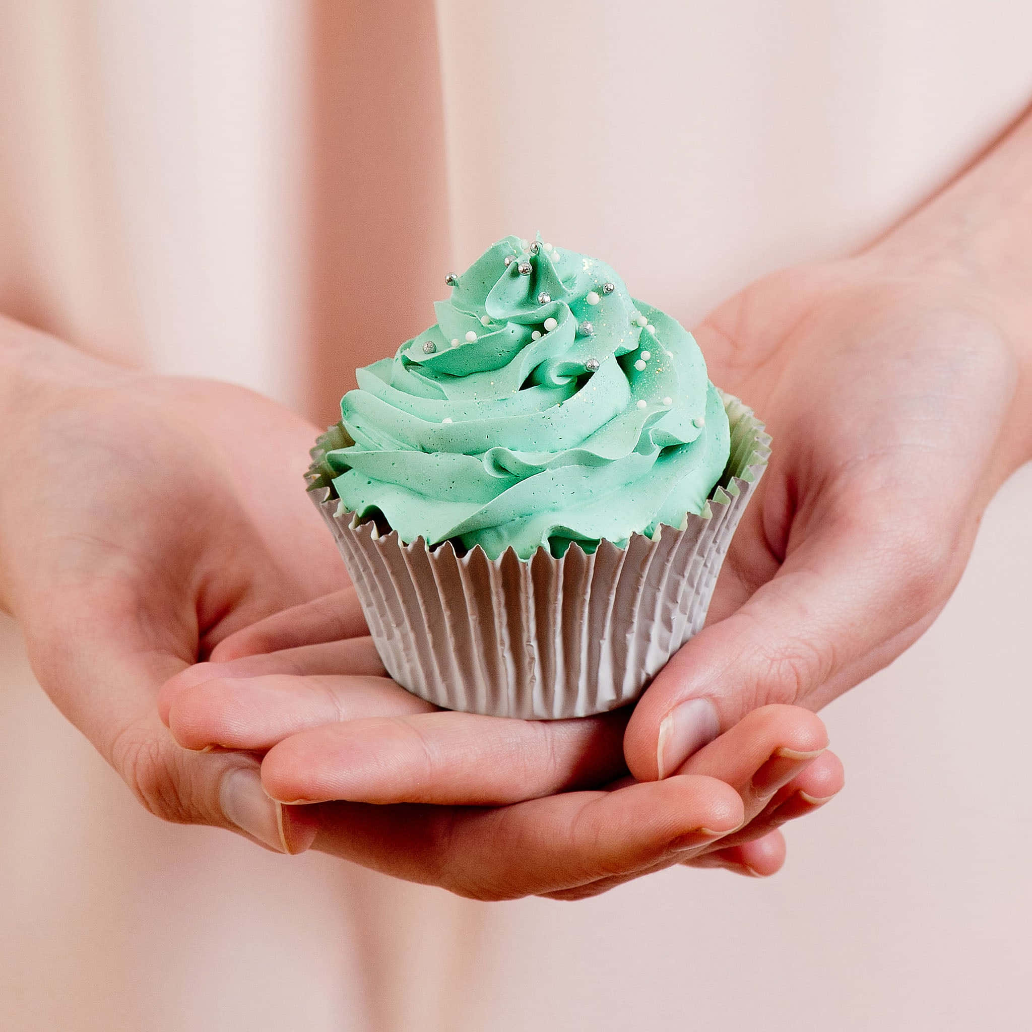 Cute Cupcake 桌布 壁紙 — 兩隻手輕輕捧著一個薄荷綠色糖霜、綴有銀色珍珠糖粒的杯子蛋糕，背景是柔和的粉紅色。