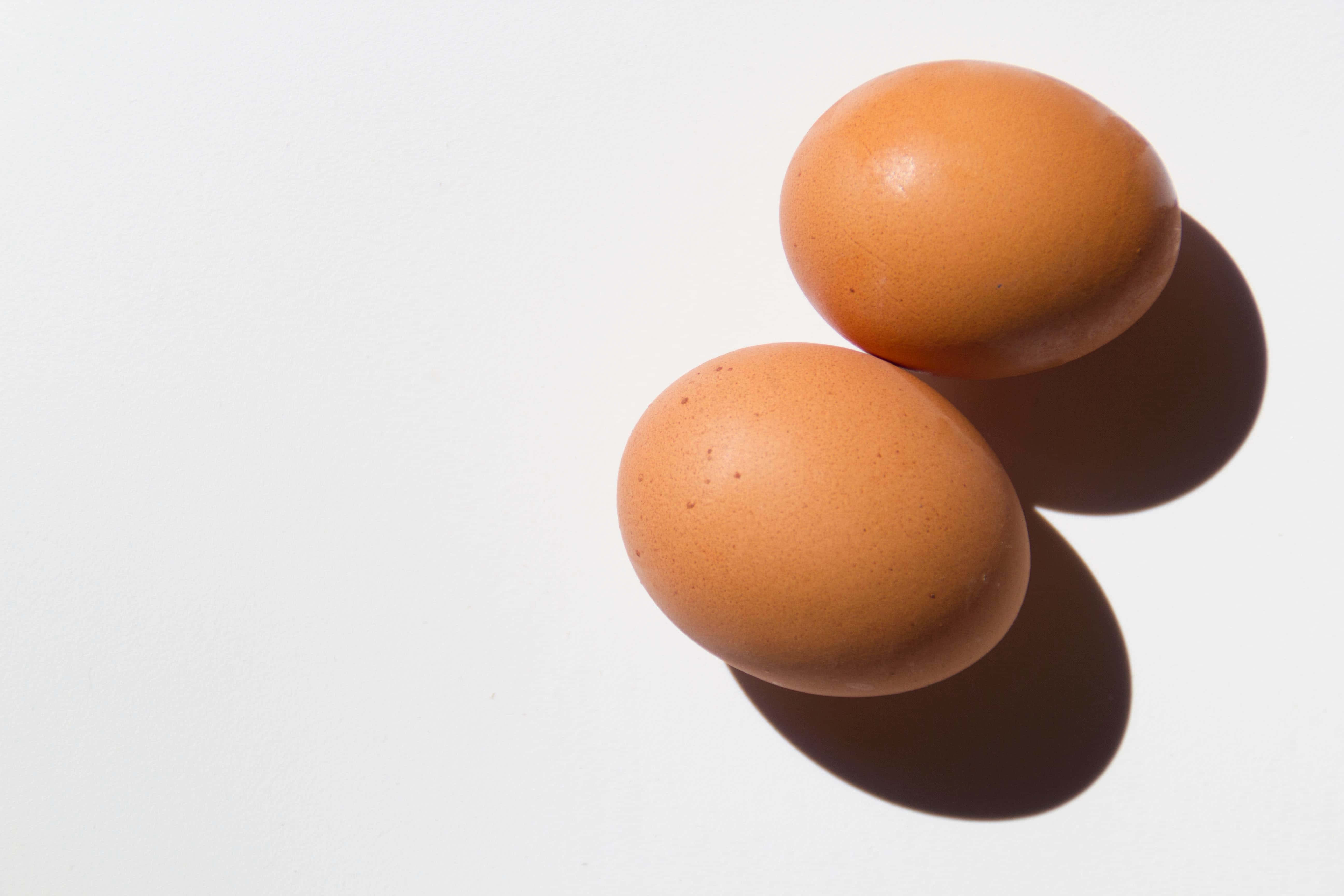 Brown Eggs 桌布 壁紙 — 兩隻棕色雞蛋放在白色表面上，在明亮光線下投射出柔和陰影。