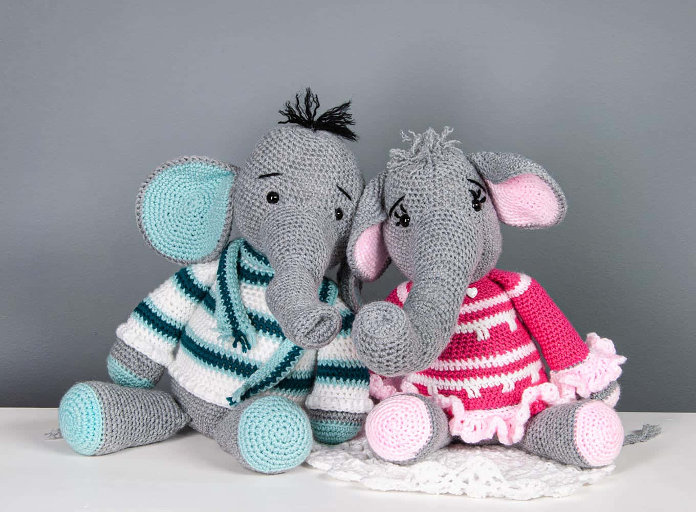 Emily Elephant 桌布 壁紙 — 兩隻穿著彩色條紋服裝的鉤針編織大象並排坐在灰色背景上的蕾絲墊上。