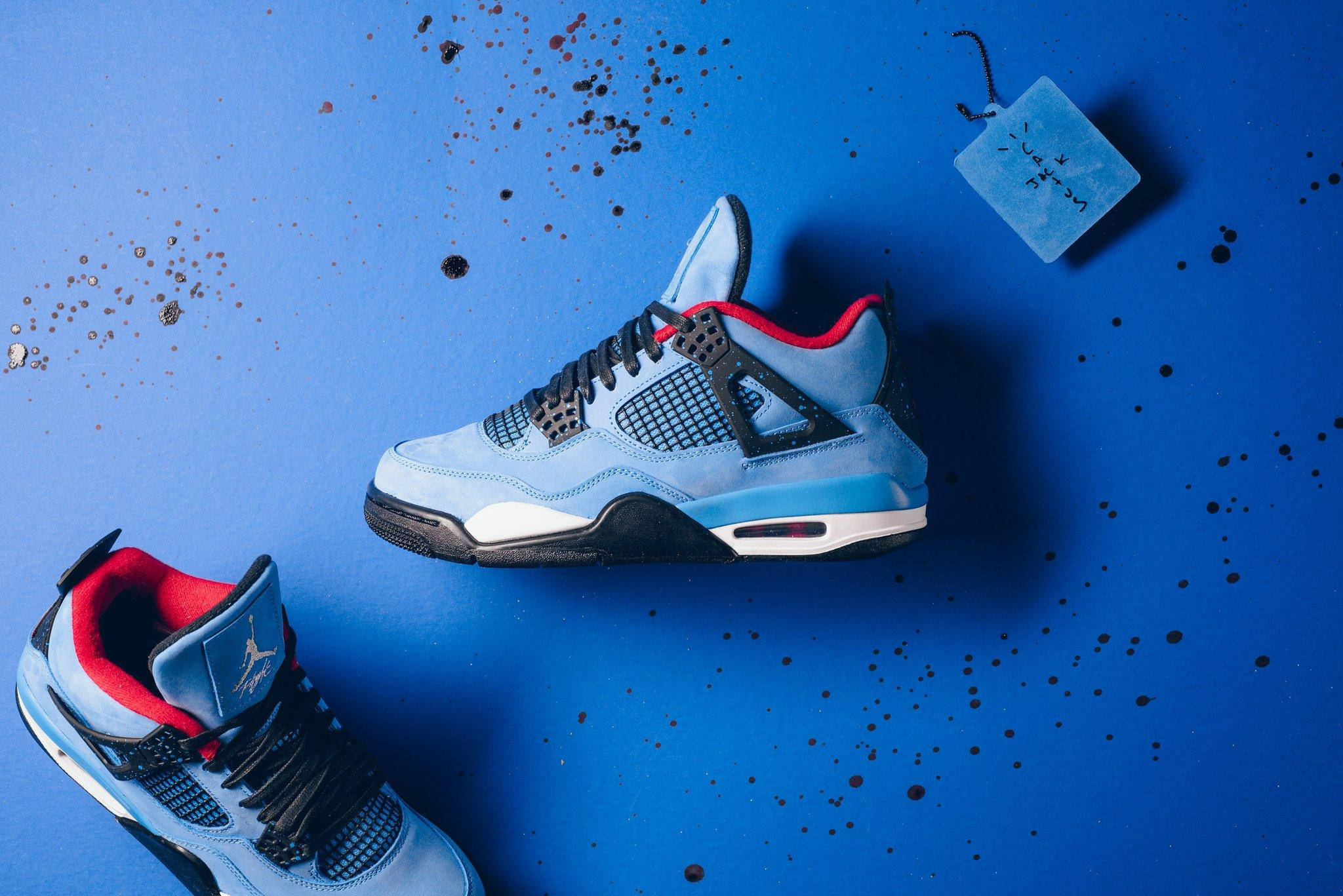 Cactus Jack Air Jordan Retro 壁紙 — 兩雙淺藍色 Jordan 4 運動鞋帶有紅色點綴，放置在濺有黑色油漆滴的鮮豔藍色表面上。