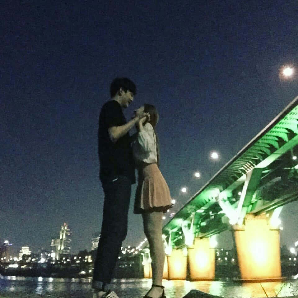 兩顆心，幸福滿溢 壁紙 — 一對情侶在夜晚發光的綠色橋下分享親密時刻，身後的城市燈光倒映在水面上。