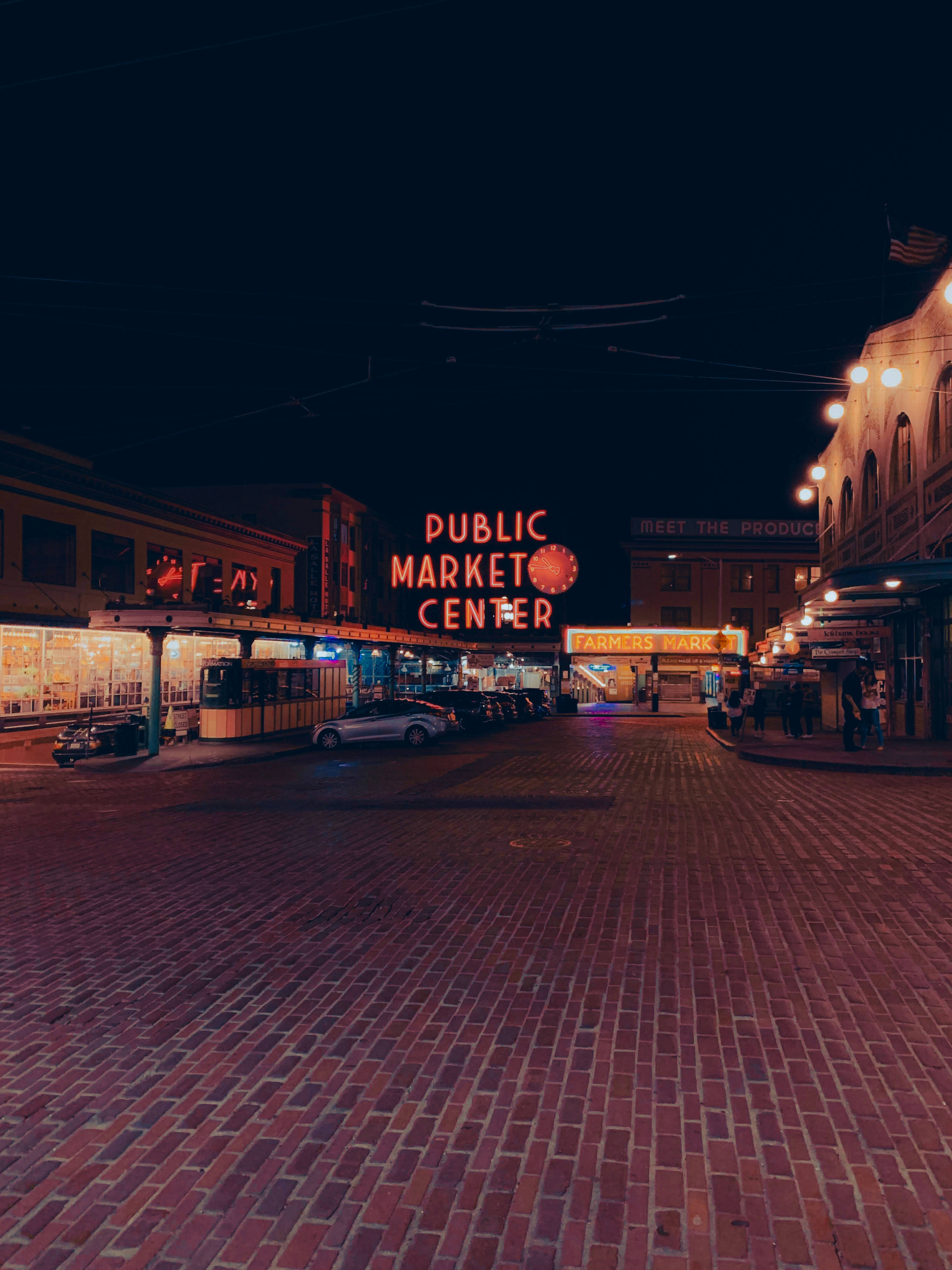 Seattle iPhone Public Market Center 壁紙 — 公共市場中心充滿活力的夜景，發光的霓虹燈照亮了鋪滿磚塊的街道，兩旁是歷史建築。