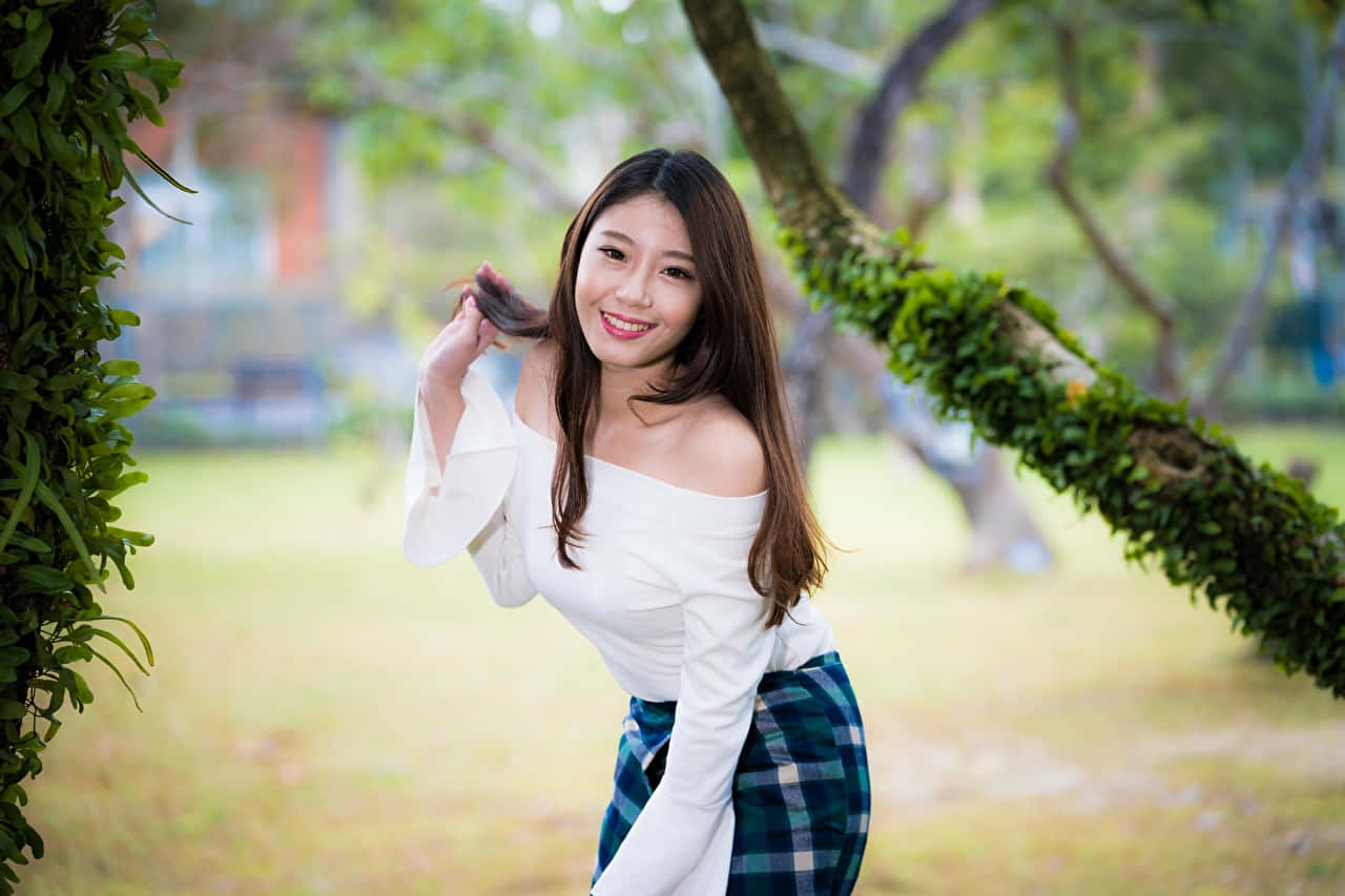 公園中的可愛亞洲女性 壁紙 — 一位微笑的年輕女性身穿白色露肩上衣和格子裙，在陽光明媚的公園裡，在長滿苔蘚的樹枝下俏皮地擺姿勢。