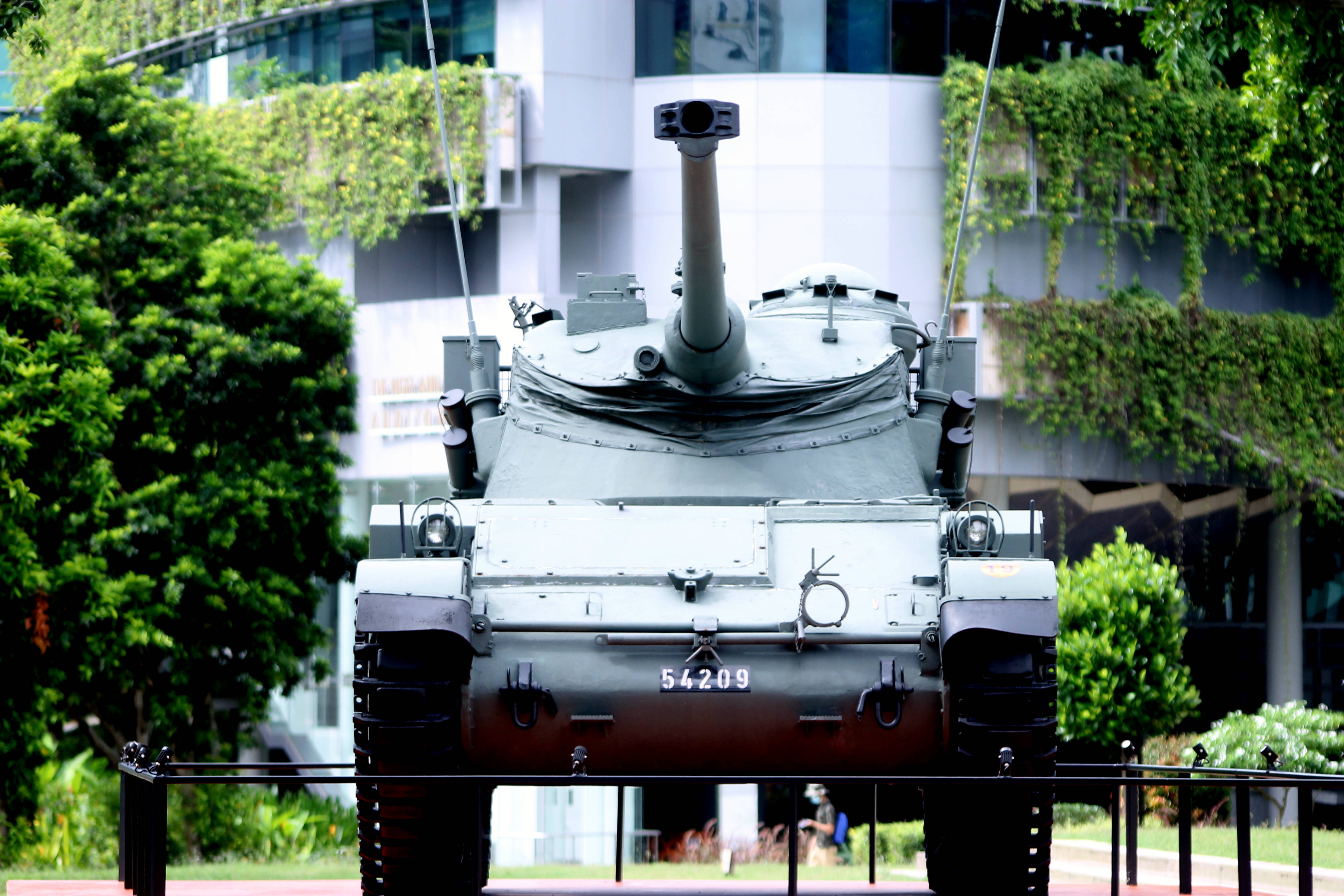 公園中的坦克紀念碑 壁紙 — 一輛灰色軍用坦克編號54209，在一個綠意盎然的城市公園中展出，周圍環繞著現代建築。