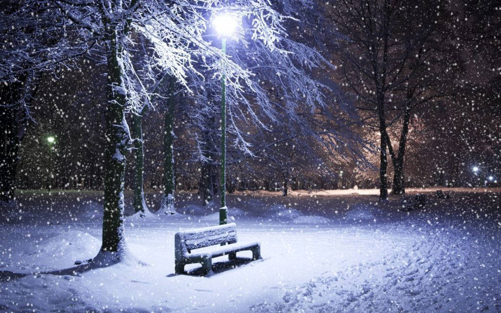 公園長椅冬季桌面壁紙 壁紙 — 一張被雪覆蓋的公園長椅在街燈下發光，夜間雪花輕輕飄過光禿的冬季樹木。