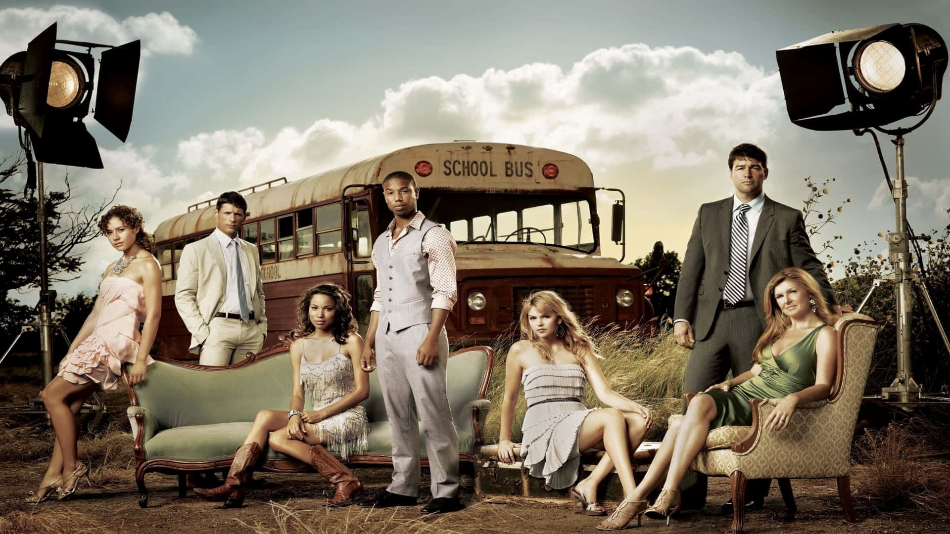 Tv Series 桌布 壁紙 — 六位衣著優雅嘅人喺田野嘅工作室燈光下，圍住復古傢俬同一架生鏽嘅校車戲劇性地擺甫士。
