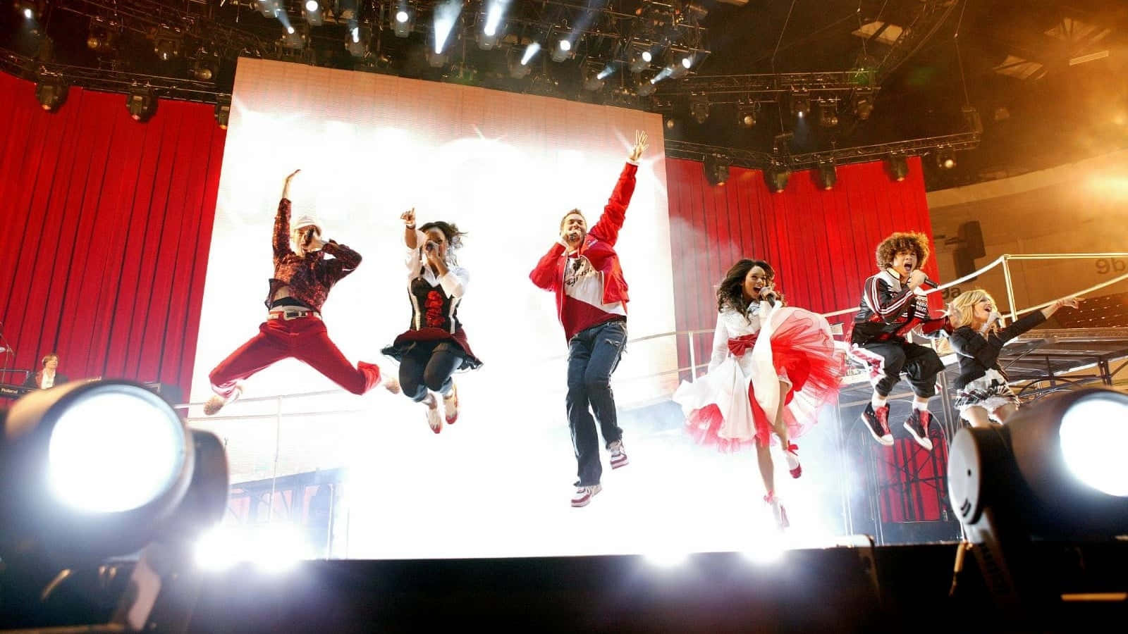 High School Musical 桌布 壁紙 — 六位表演者在紅色帷幕和聚光燈框住的明亮舞台上充滿活力地跳躍。