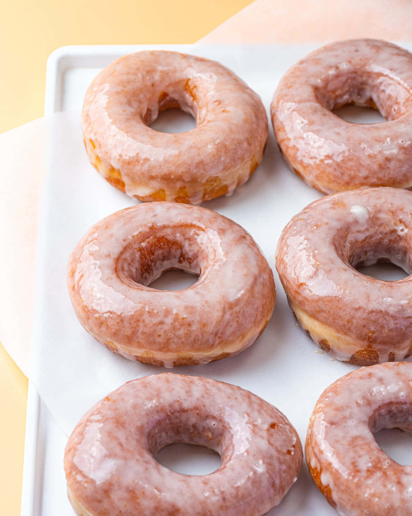 Glazed Donut 桌布 壁紙 — 六個帶有閃亮粉紅色糖霜的釉面冬甩整齊地放在白色托盤上，背景是柔和的黃色。