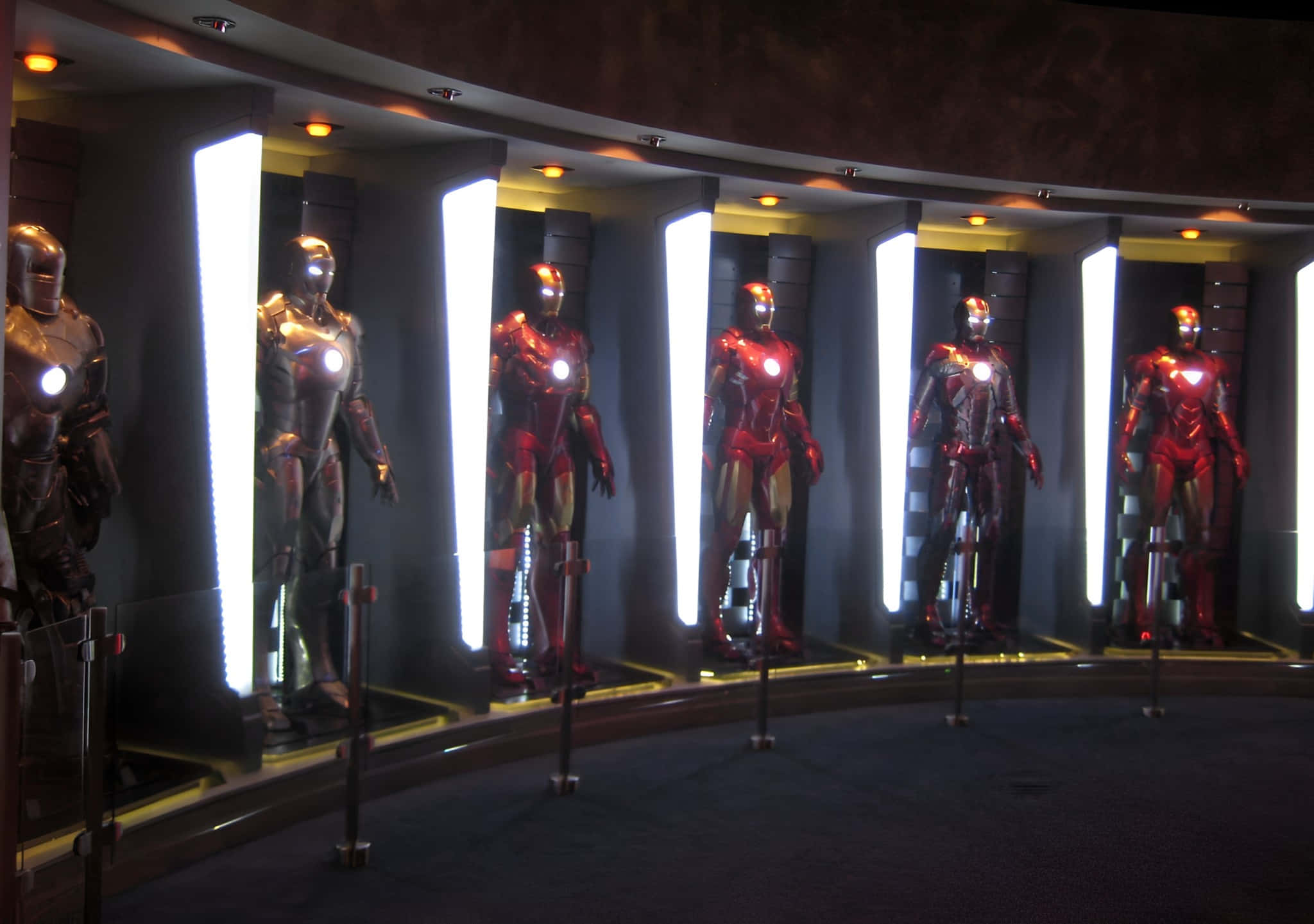 Iron Man Suit 桌布 壁紙 — 六個發光的鐵甲奇俠套裝在昏暗的博物館環境中，在明亮的垂直燈光下，站立在彎曲的展示櫃中。