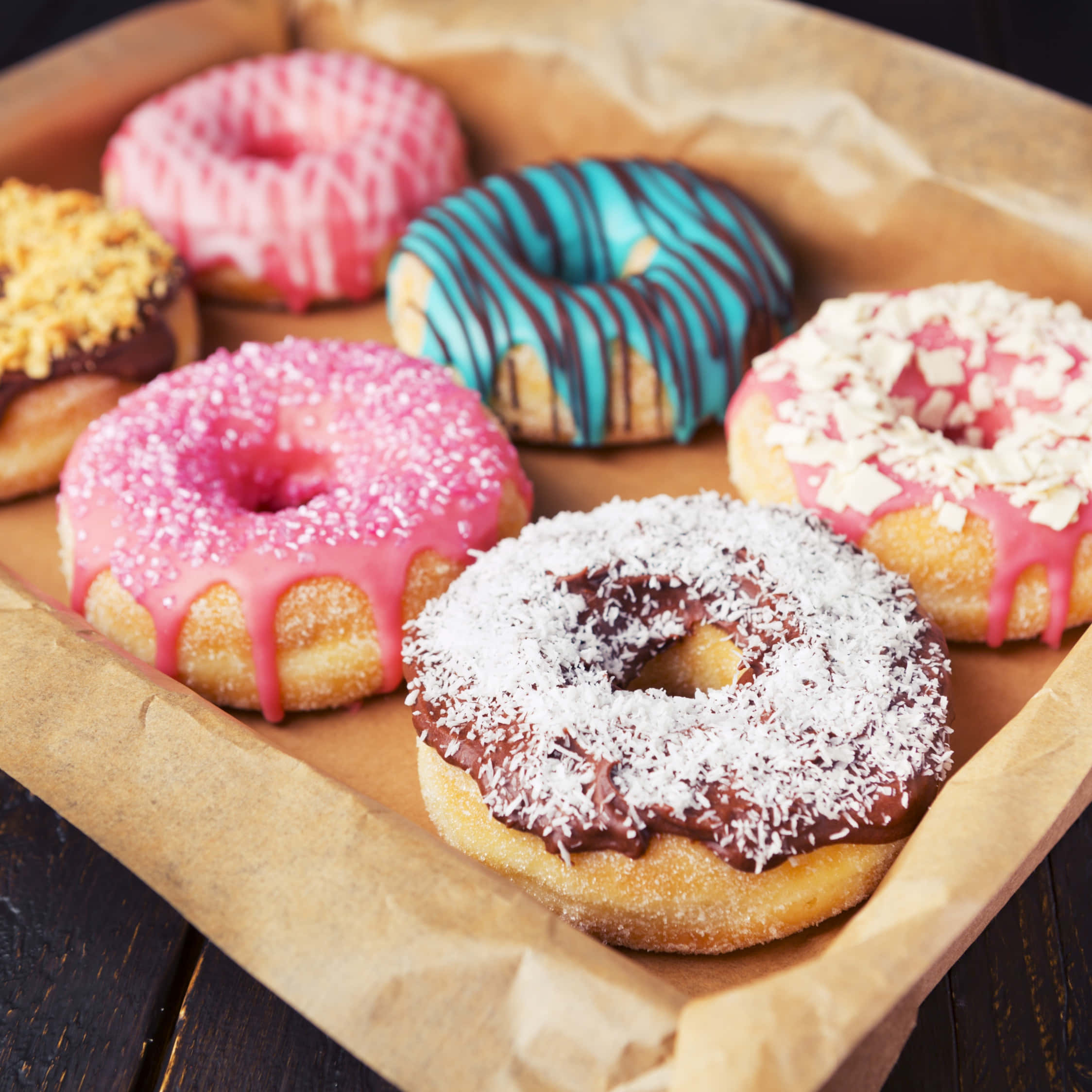 Cute Donut 桌布 壁紙 — 六個色彩鮮豔的釉面甜甜圈，上面有粉紅色、藍色、朱古力和椰子配料，放在一個鋪有棕色紙的托盤上，置於深色木質表面。