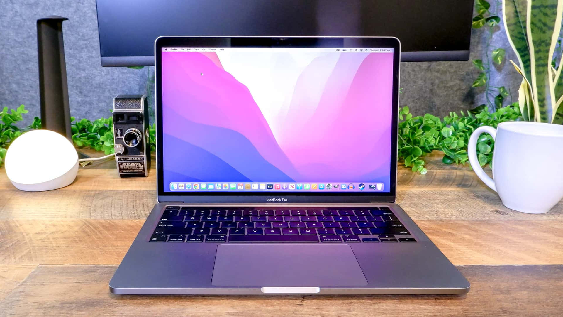 具備時尚現代設計的MacBook筆記本電腦 壁紙 — 一部時尚的銀色MacBook Pro打開放在木桌上，周圍有綠色植物、白色杯子和復古相機。