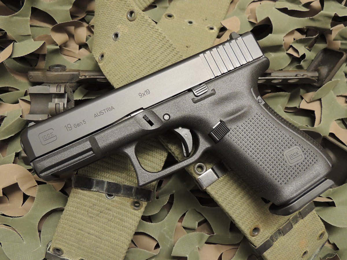 具有磨砂黑色塗裝的 Glock 19 手槍 壁紙 — 一把黑色Glock 19 Gen5手槍放在橄欖綠色迷彩布料上，布料有明顯磨損和縫線。