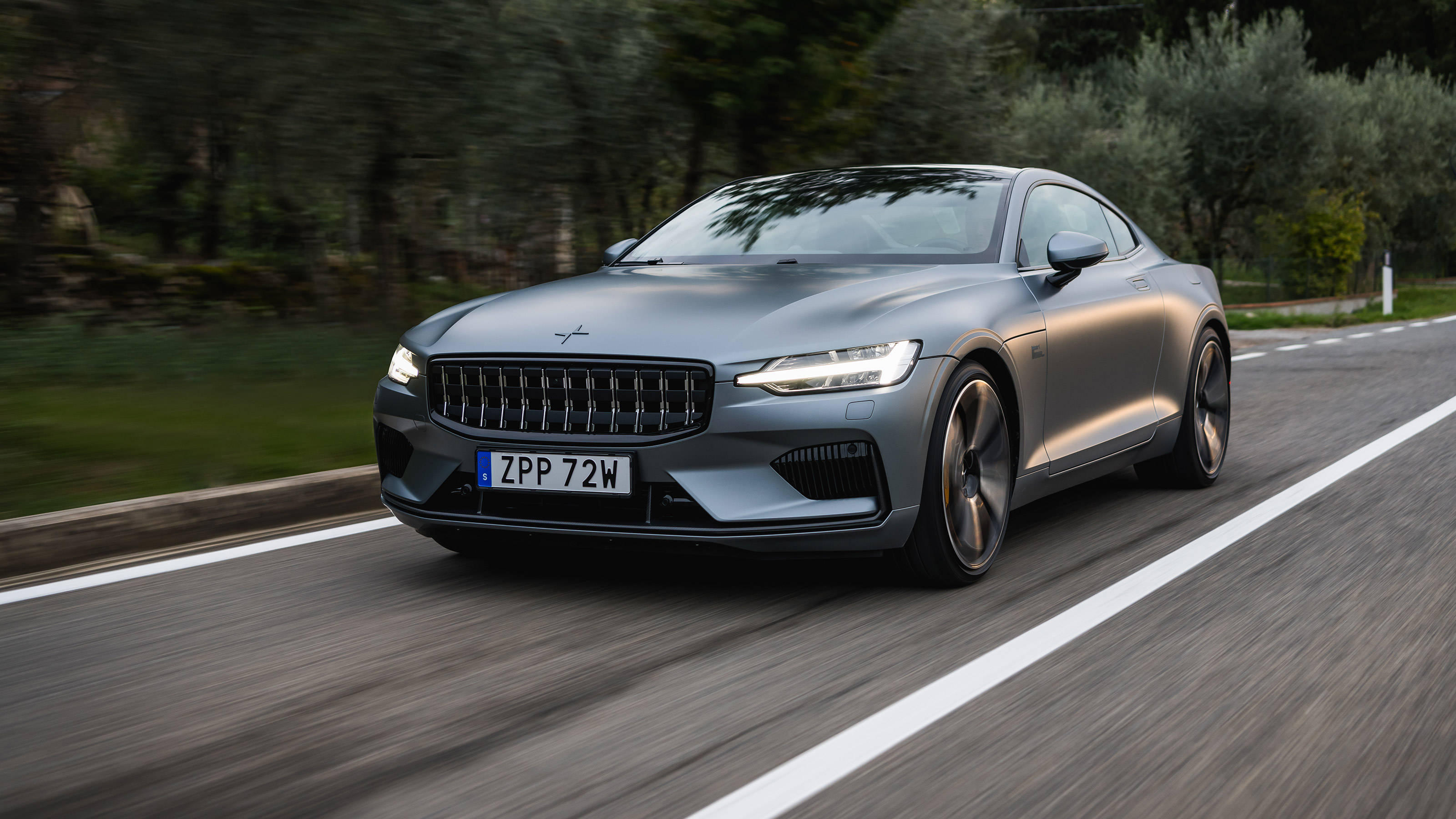 具流線設計的Polestar 1 壁紙 — 一輛時尚的灰色Polestar 2跑車沿著蜿蜒的道路穿過茂密的綠色森林飛馳。