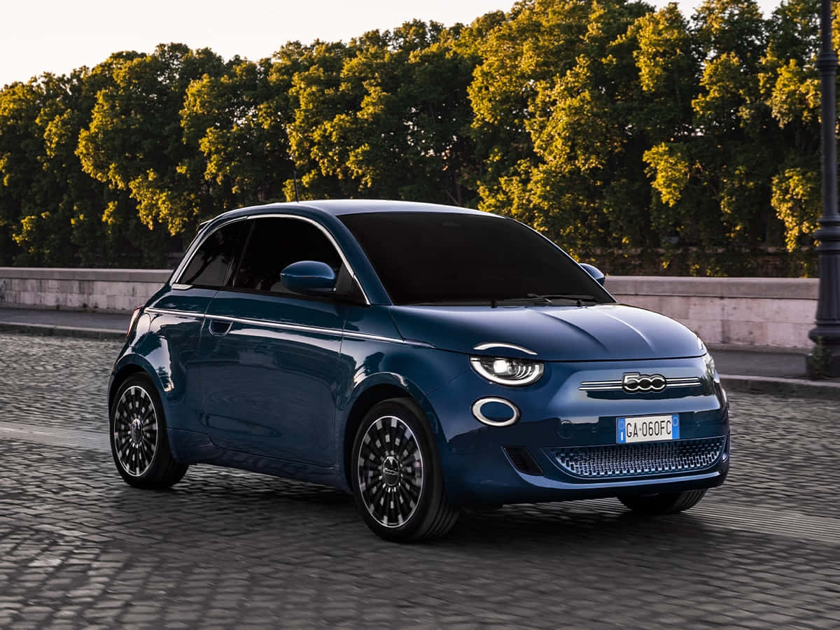 典雅義大利菲亞特 壁紙 — 一輛時尚的藍色電動Fiat 500e在鵝卵石街道上行駛，街道兩旁綠樹成蔭，沐浴在金色的夕陽下。