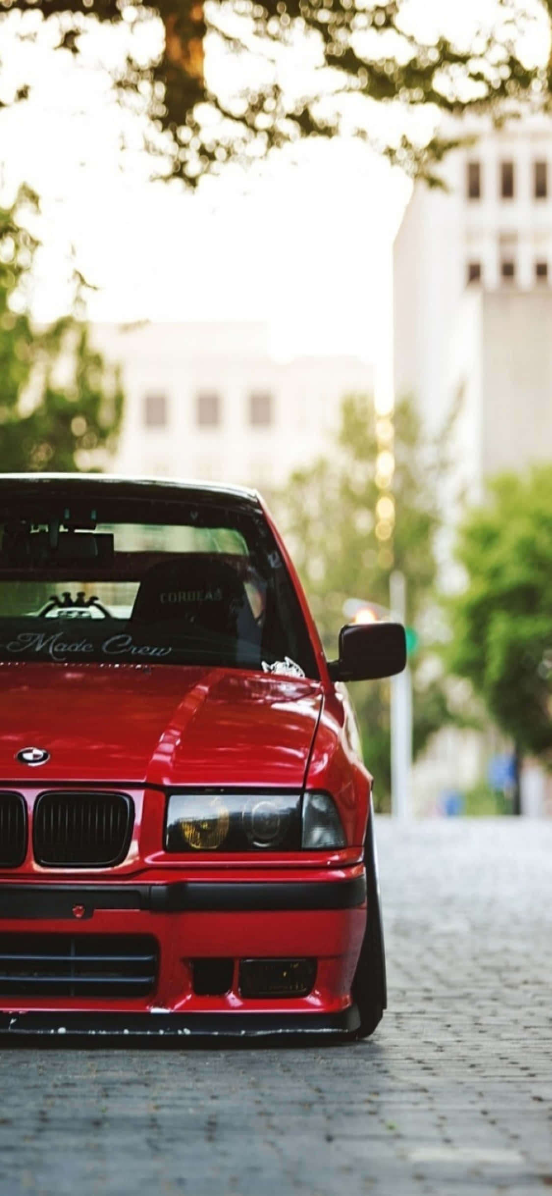 典雅與實用的完美結合：iPhone X與BMW 壁紙 — 一輛光亮的紅色BMW E30跑車停在鵝卵石街道上，斑駁的陽光下，背景有模糊的綠色樹木和建築物。