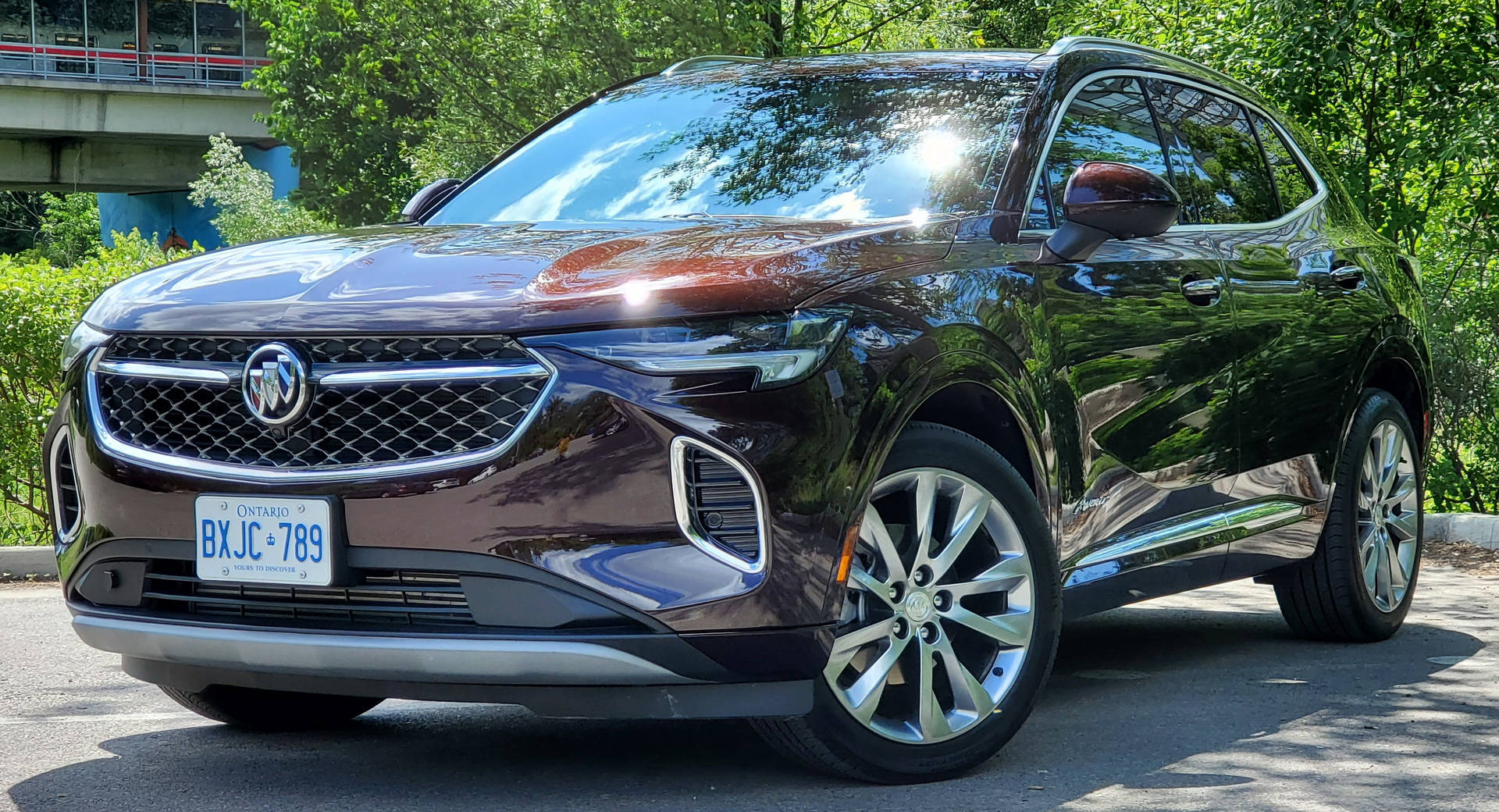 栗色Buick SUV在陽光明媚的日子停車，背景有綠樹和橋樑。