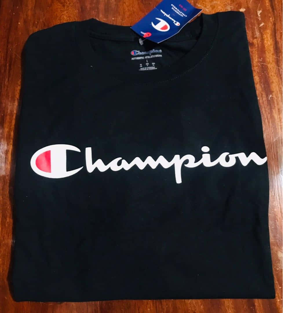 冠軍：以自信邁向更高 壁紙 — 一件整齊折疊的黑色Champion T恤，帶有白色標誌，放在溫暖的木質表面上。