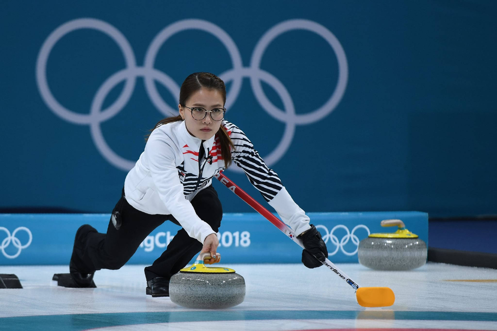 冬季奧運女子冰壺運動員 壁紙 — 一位戴眼鏡、專注的女性冰壺運動員在冰面上推動花崗岩冰壺，身後可見奧運五環。