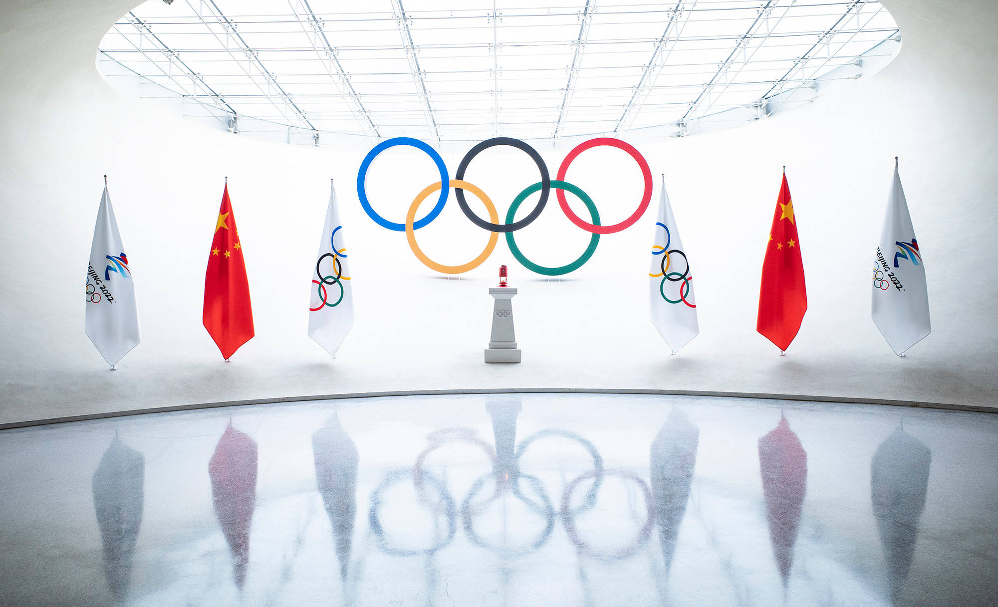 冬季奧運會高速下坡滑雪選手 壁紙 — 奧運五環懸掛在明亮的白色圓頂內，中國國旗和奧運旗幟反射在拋光地板上。