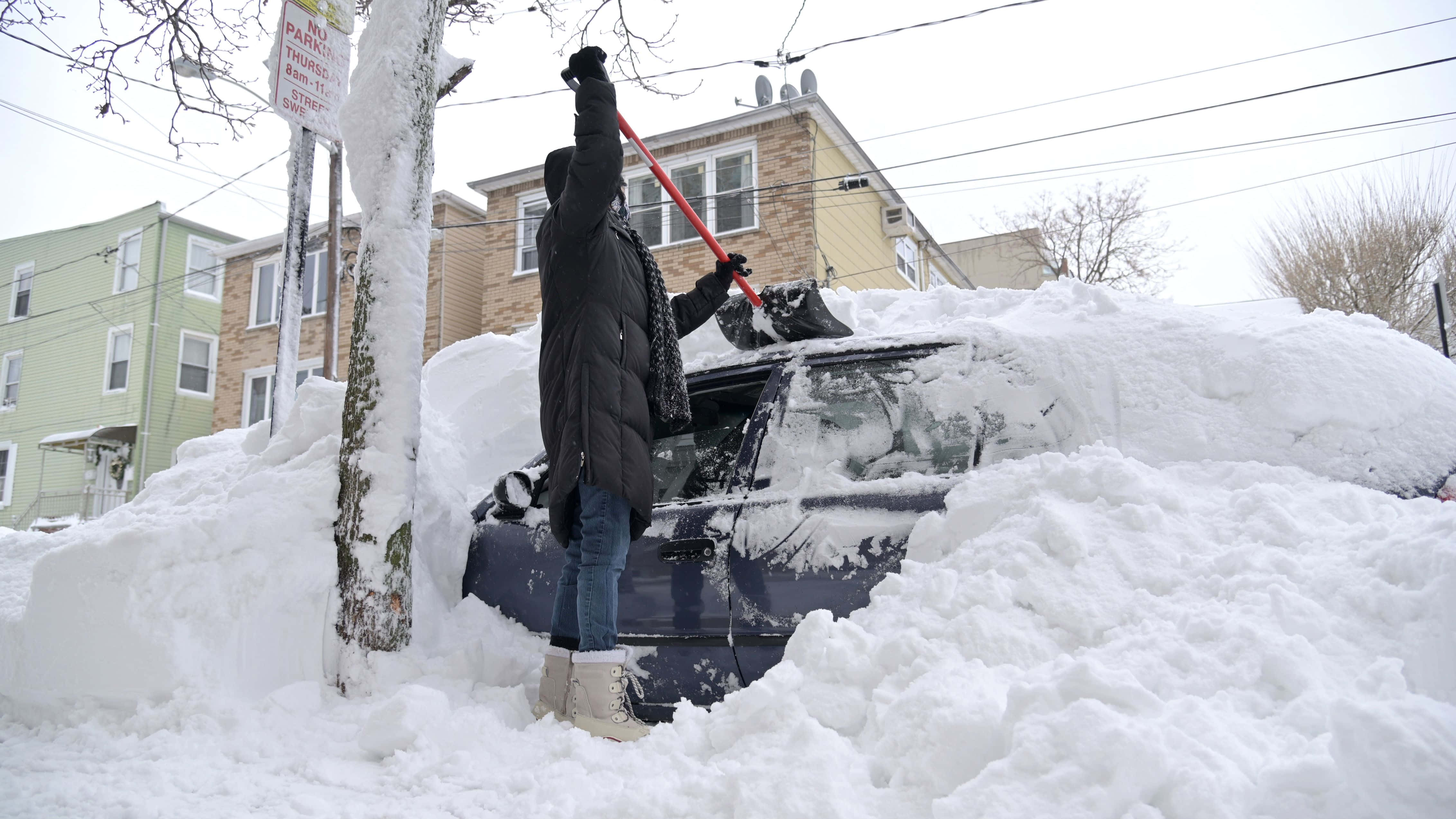 冬季暴雪帶來雪花與涼爽氣溫 壁紙 — 一名身穿黑色外套的人正在住宅區清理一輛被厚厚白雪覆蓋的汽車上的積雪。