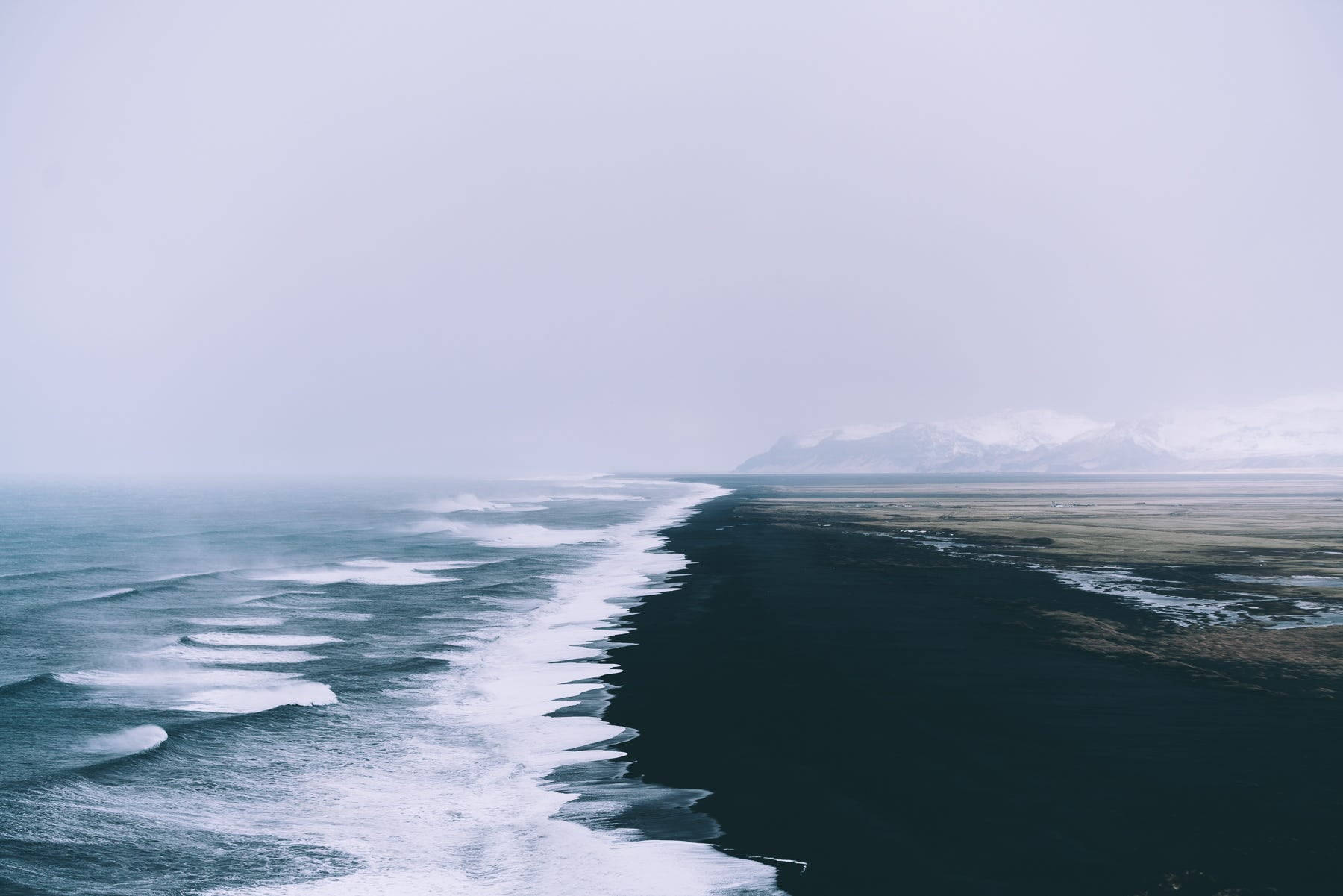 冬季海景 壁紙 — 黑色沙灘的清晰鳥瞰圖，海浪拍打著霧氣瀰漫的地平線，遠處有積雪的山峰。
