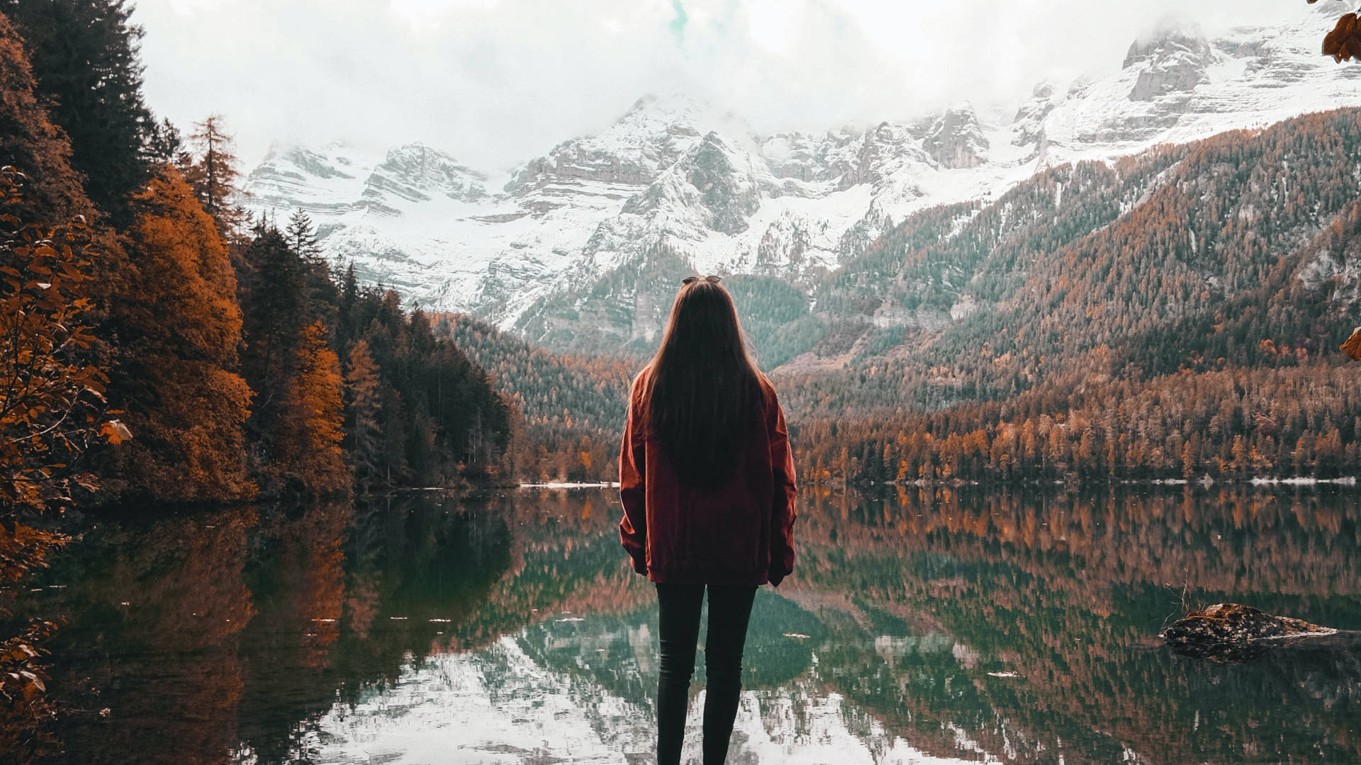 穿紅色外套的人站在湖邊，湖面倒映山巒