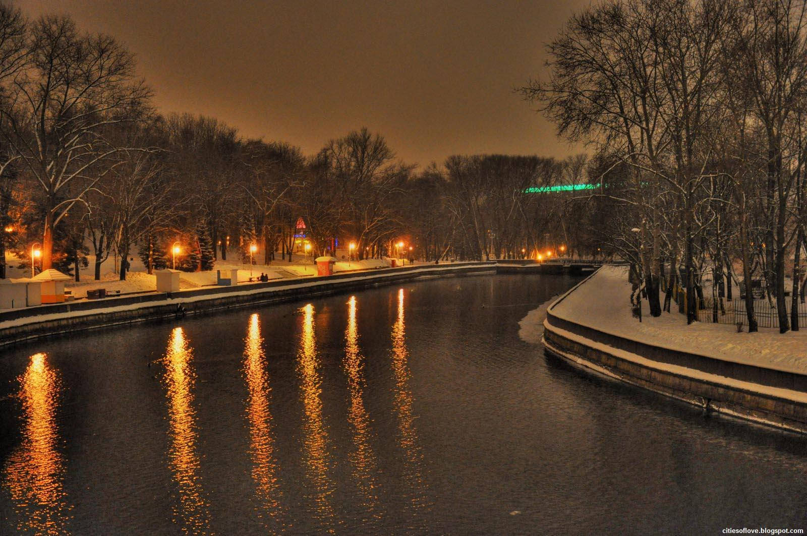 冬季白俄羅斯河畔夜景 壁紙 — 一個雪白的冬夜，沿著平靜的運河，溫暖的路燈倒映在水面上，光禿禿的樹木排列在兩岸。