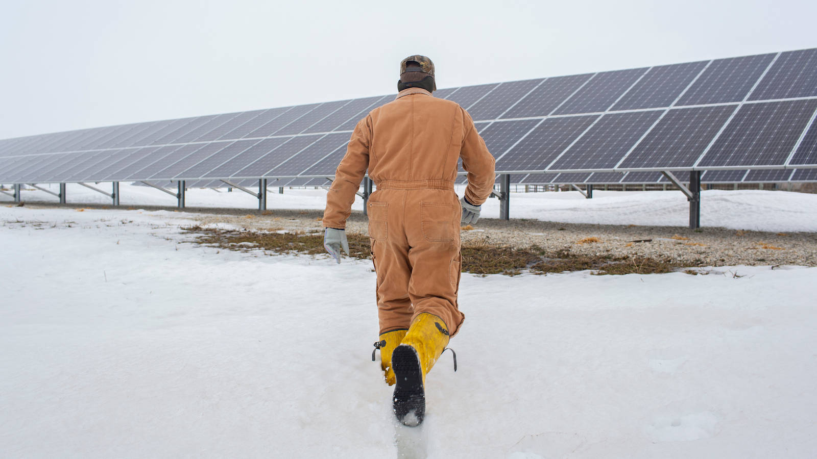 冬季的NV Energy太陽能公園 壁紙 — 一名身穿橙色連身工作服的工人，在陰天之下穿過雪地走向一排排太陽能板。
