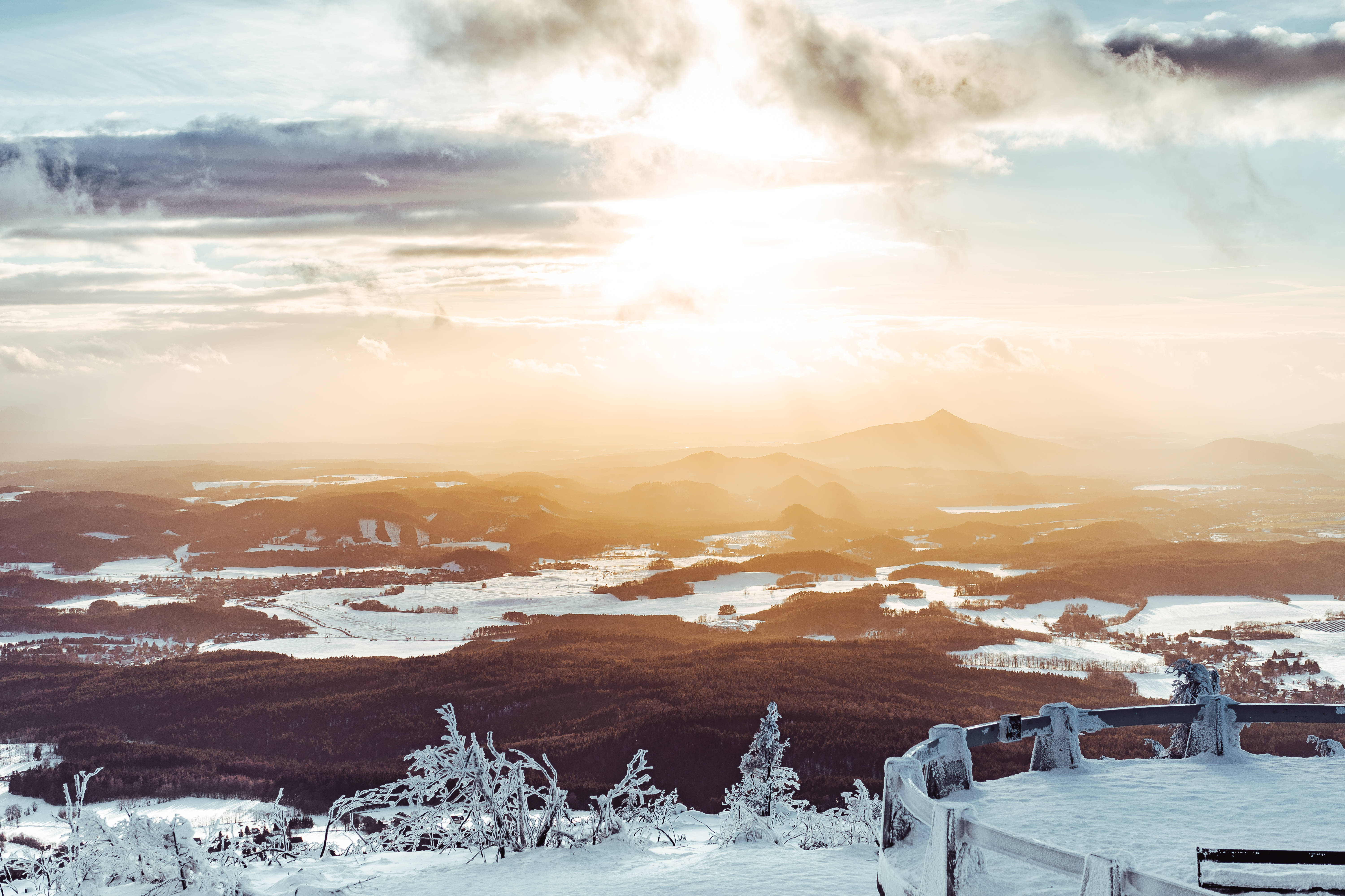 冬後自然風景 壁紙 — 夕陽將白雪覆蓋的山谷沐浴在金色光芒中，霜凍的樹木和遠山在陰雲密布的天空下。
