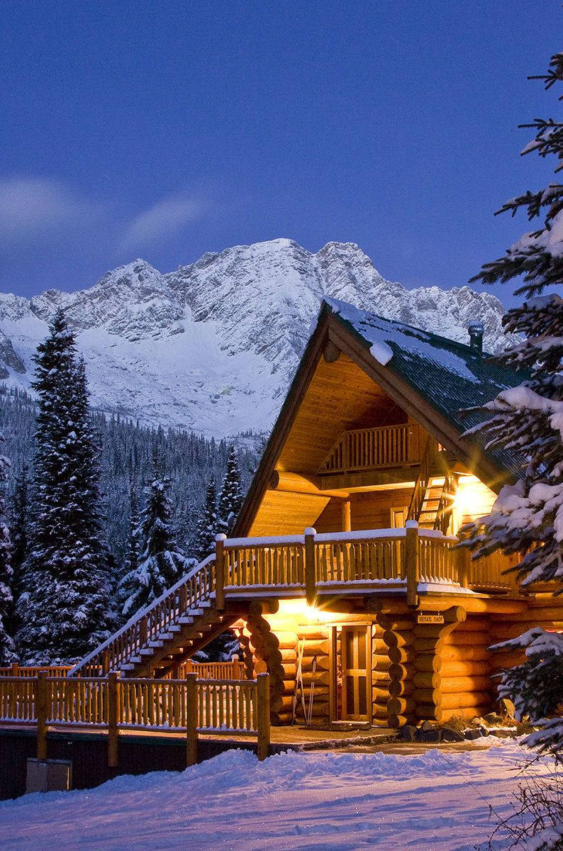 冬日小屋與晴空 壁紙 — 在深藍色暮色天空下，一座舒適的雪覆蓋小木屋在雄偉的雪山背景下溫暖地發光。