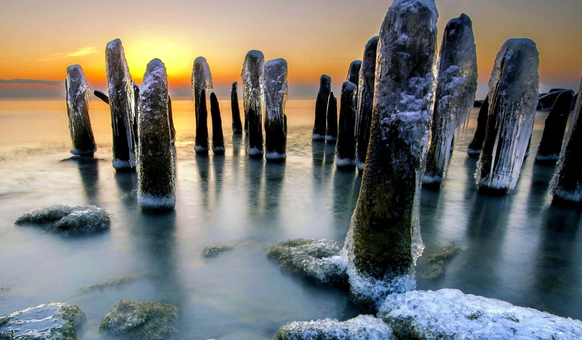 Frozen Lake 桌布 壁紙 — 冰凍的木樁豎立在平靜的冰冷水中，金色的夕陽在它們背後閃耀。