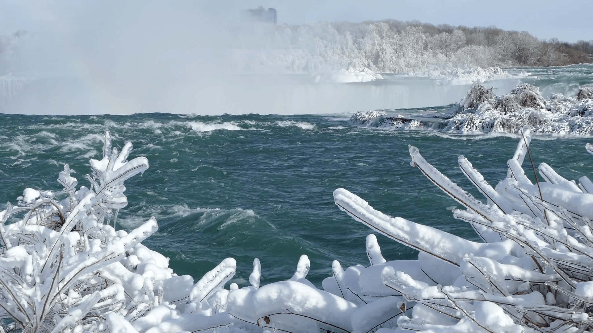 Polar Vortex 桌布 壁紙 — 冰凍的樹枝框住了一個咆哮的冬季瀑布，霧氣在清澈藍天下從冰冷的水面升起。