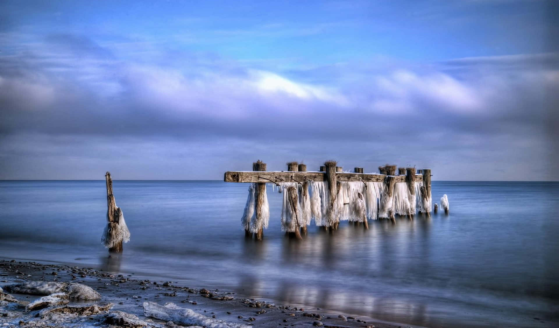 Winter Beach 桌布 壁紙 — 冰凍碼頭殘骸在平靜的水域中莊嚴地矗立，上方是帶有條紋雲朵的陰鬱藍天。
