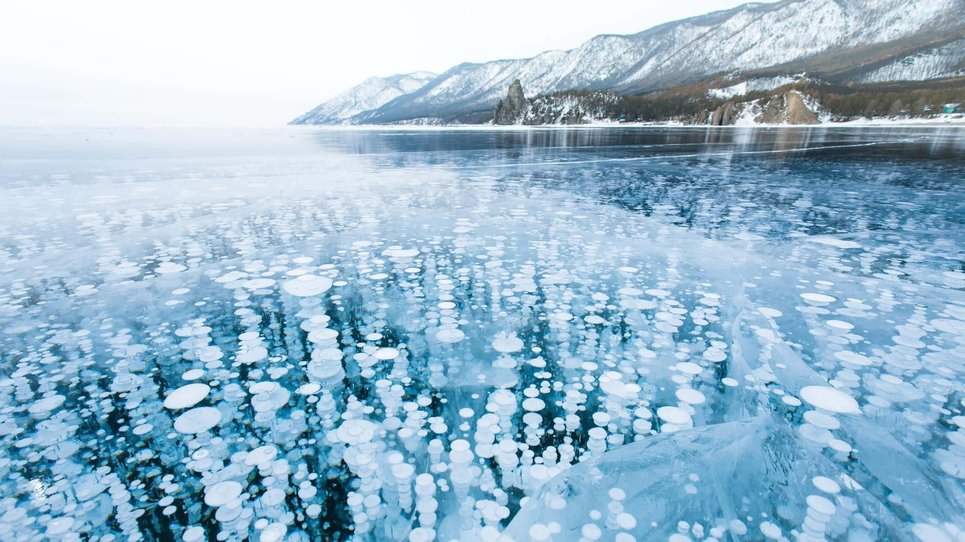 Frozen Lake 桌布 壁紙 — 冰封湖面點綴著氣泡結構，在蒼白天空下映照著積雪覆蓋的山峰。
