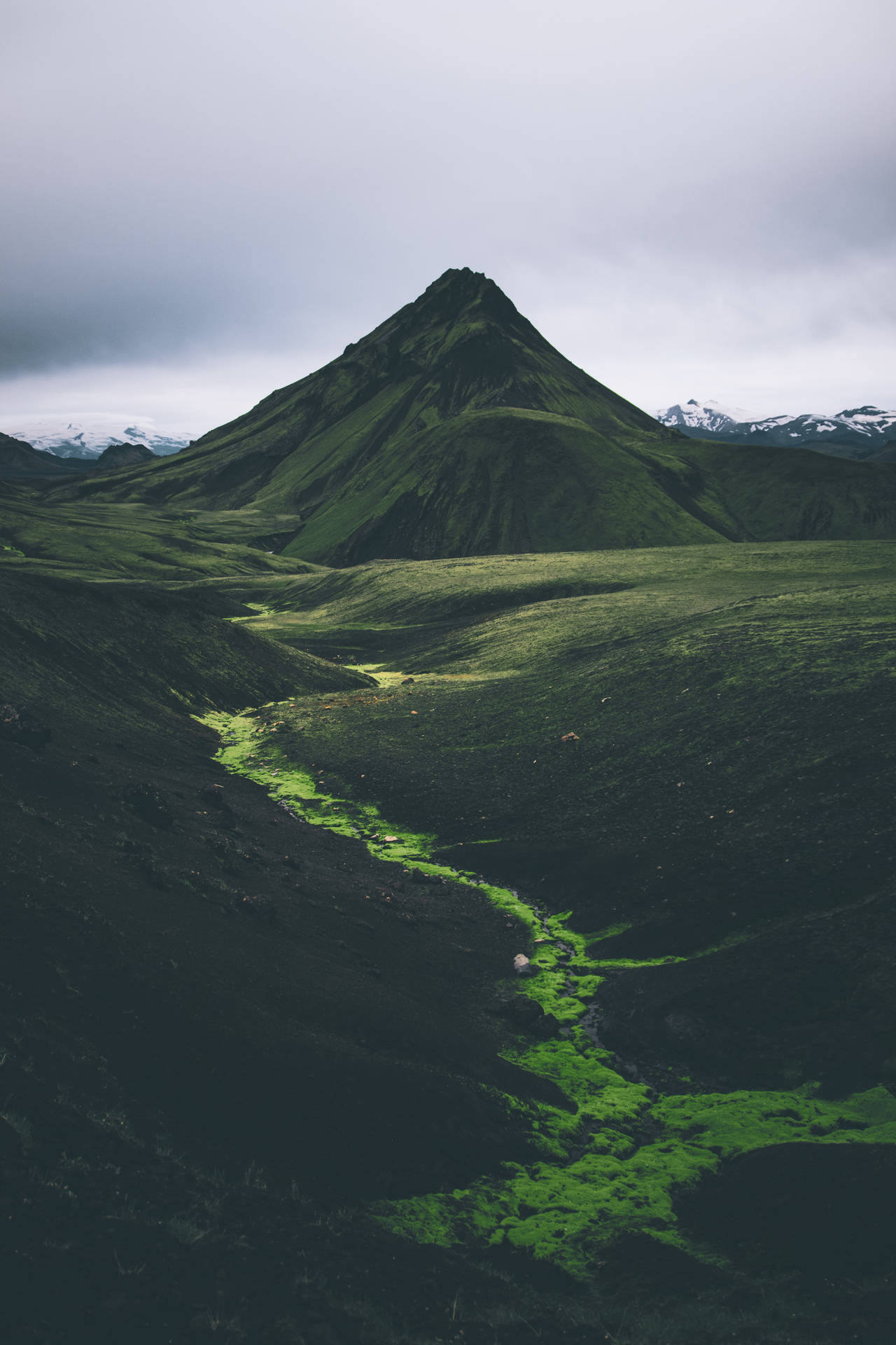 一條綠色河流流經深色火山山谷，上方是多雲的山峰。