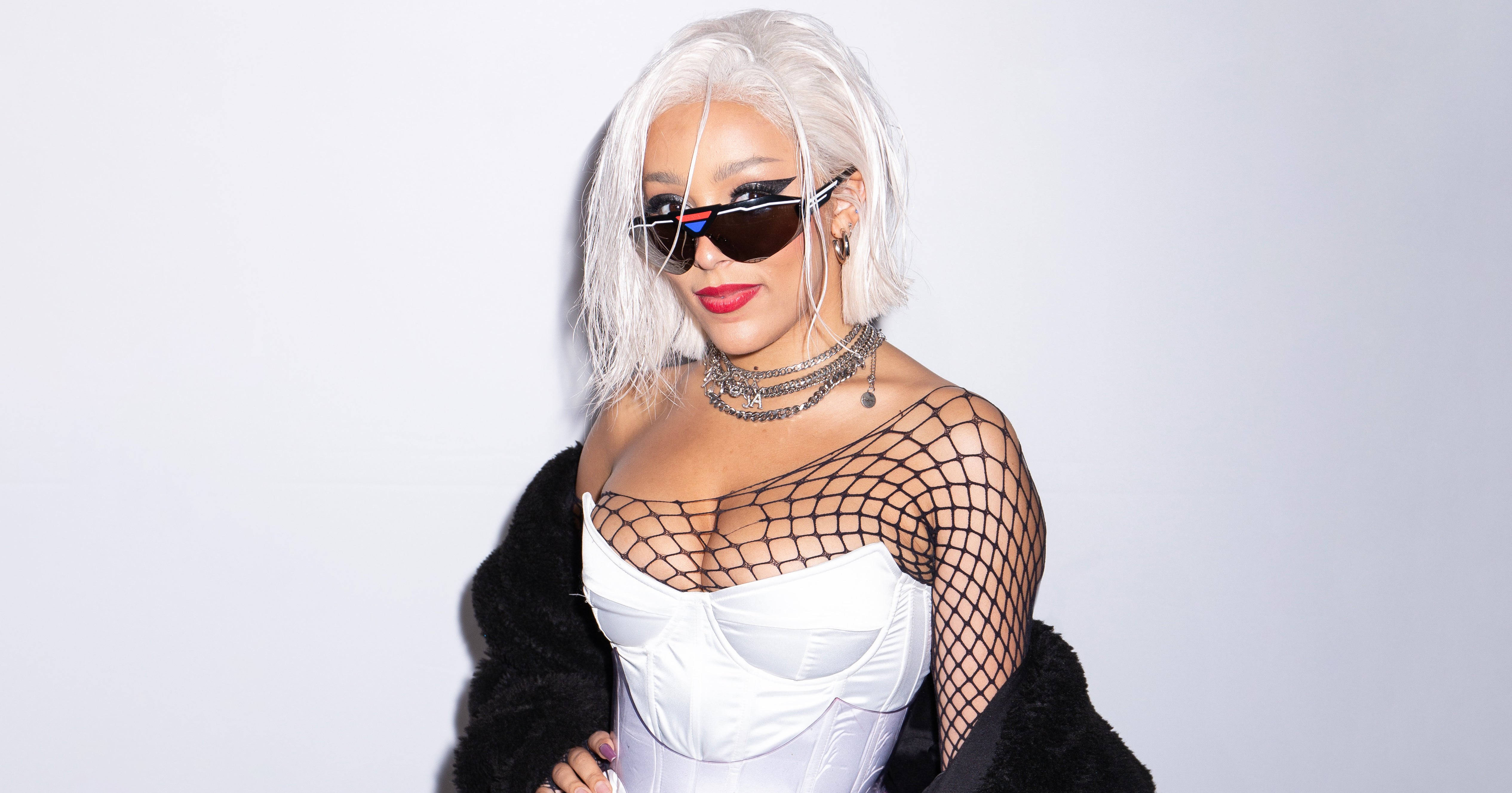 冰金髮Doja Cat 壁紙 — 一位時尚女性，擁有鉑金色頭髮和太陽眼鏡，穿著白色束身衣和黑色漁網上衣，在純白背景前自信地擺姿勢。