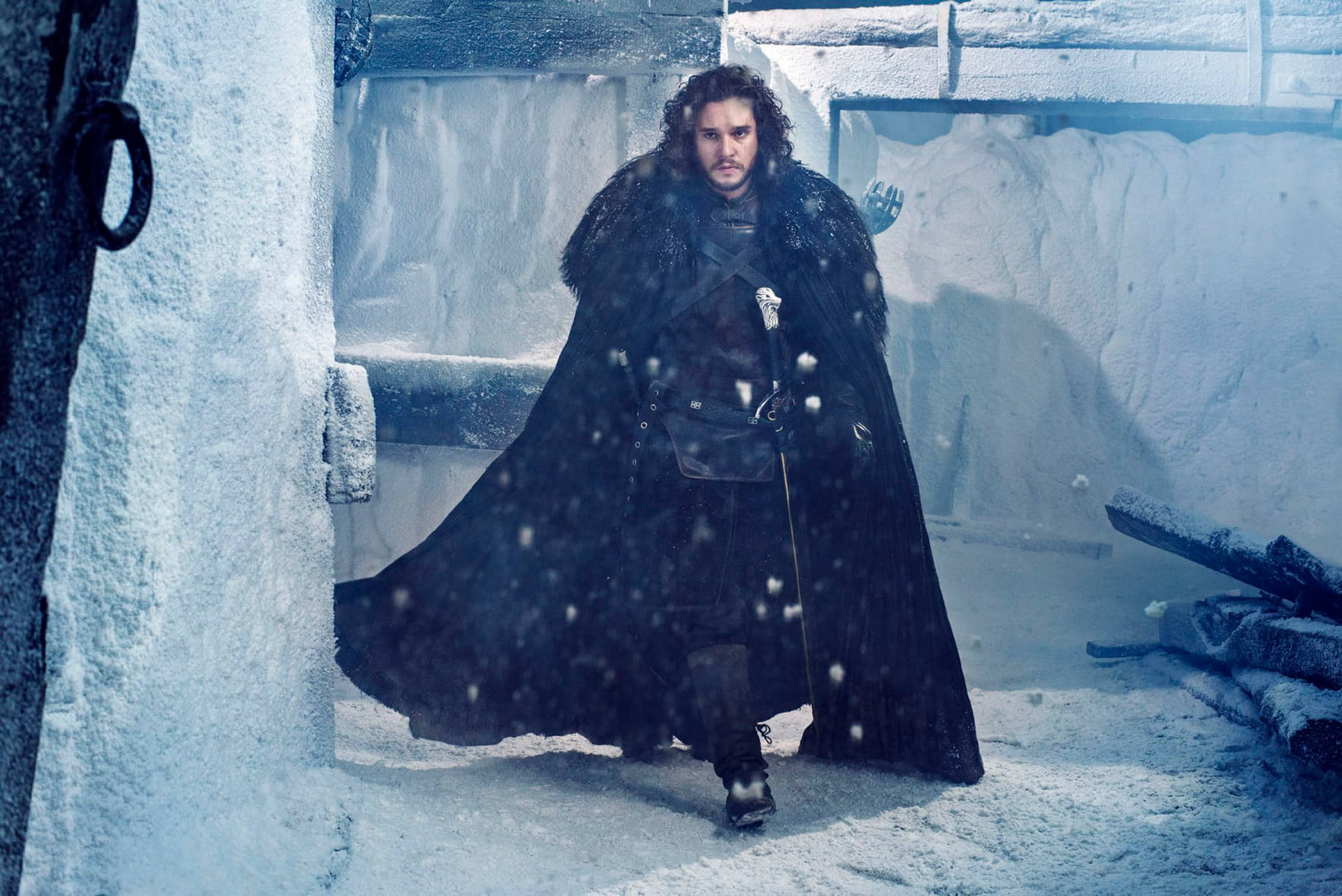 一個穿著黑色毛皮斗篷的男人在下雪時站在一個積雪的石头結構中。