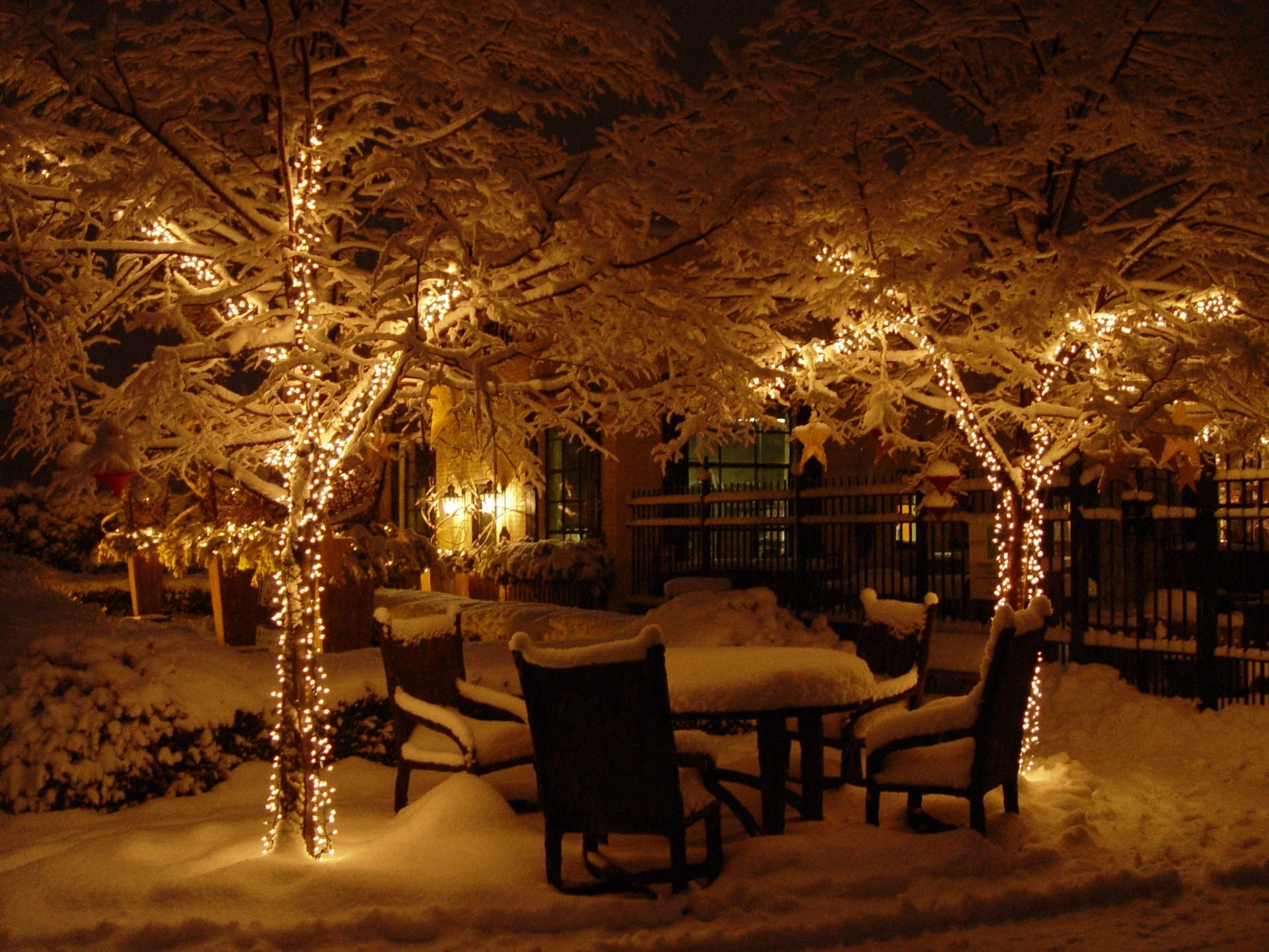 冷冬後院聖誕燈光 壁紙 — 白雪覆蓋著一個舒適的戶外庭院夜晚，溫暖的串燈在積雪的樹木上閃爍，圍繞著一張木桌和椅子。