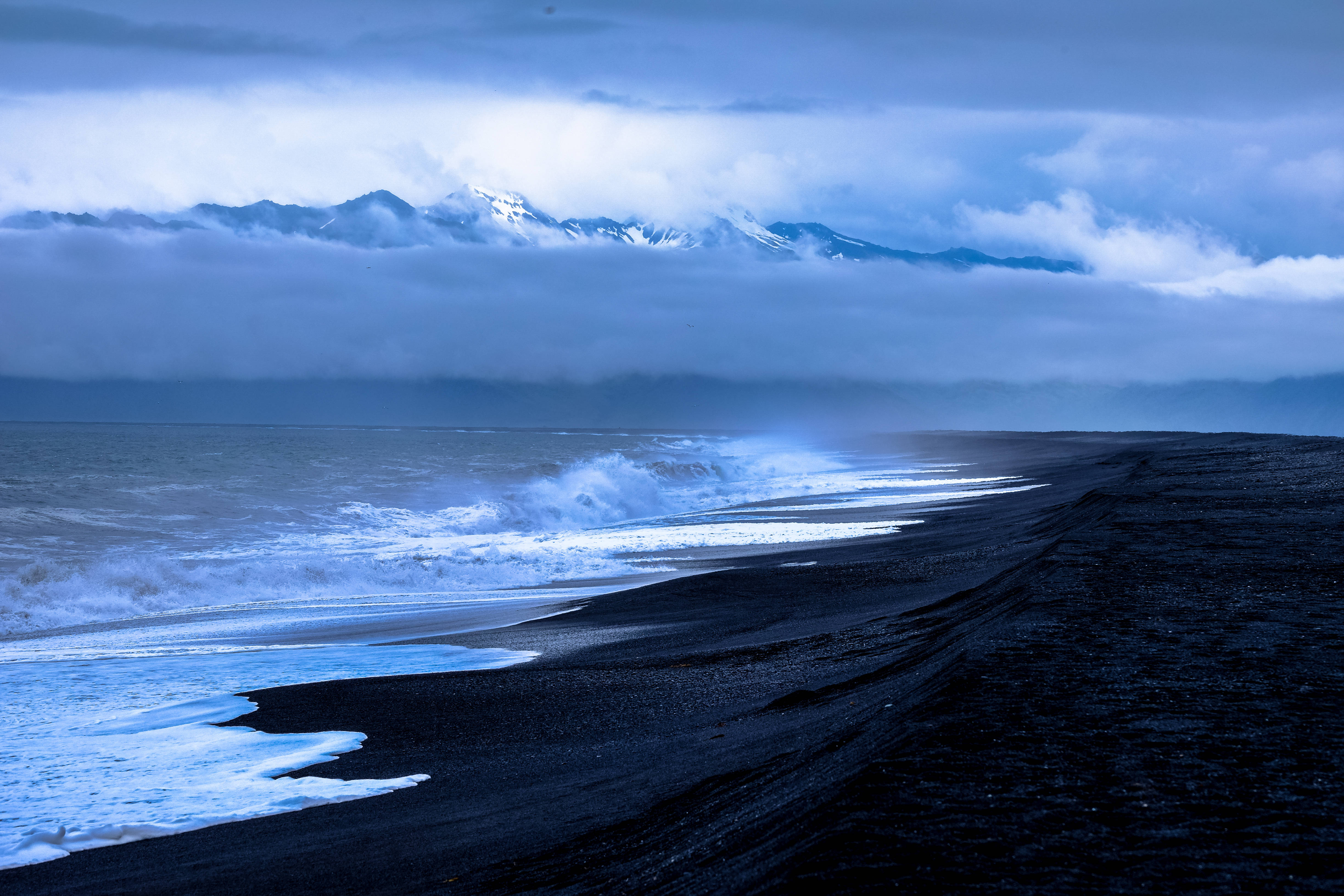 冷藍海灘 壁紙 — 一個戲劇性的海岸場景，黑色沙灘上波濤洶湧，陰沉的藍天下，積雪的山峰在雲層中若隱若現。