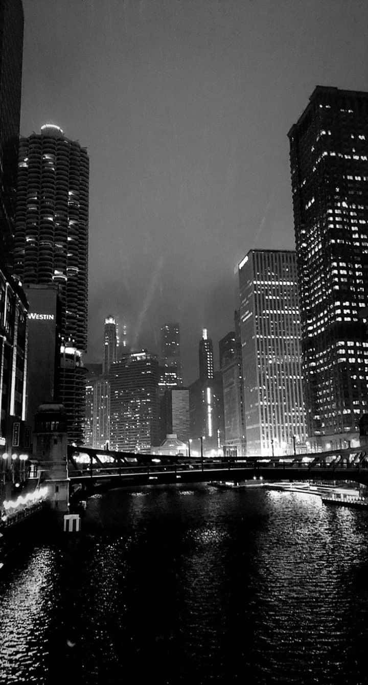 冷靜而深邃的幽暗風格背景 壁紙 — 單色夜景城市景觀，高聳的摩天大樓將燈光反射在下面的河流上。