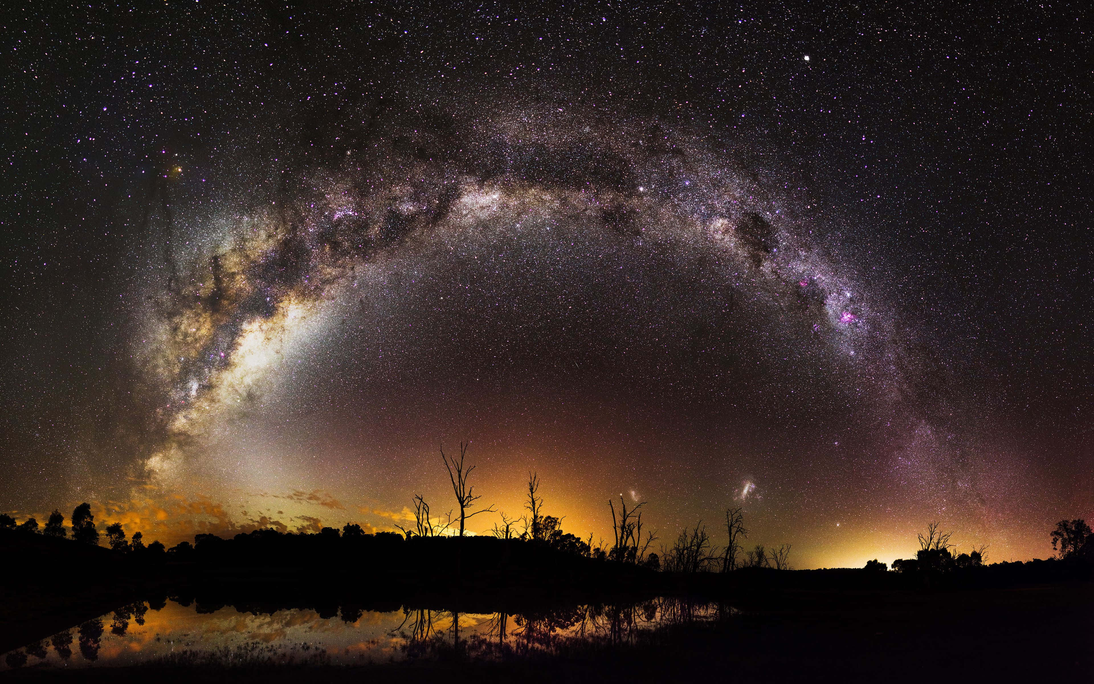 凝視繁星夜空，感受無限驚奇 壁紙 — 令人嘆為觀止的夜空在平靜的水面上拱起，倒映著銀河和發光的橙色地平線。