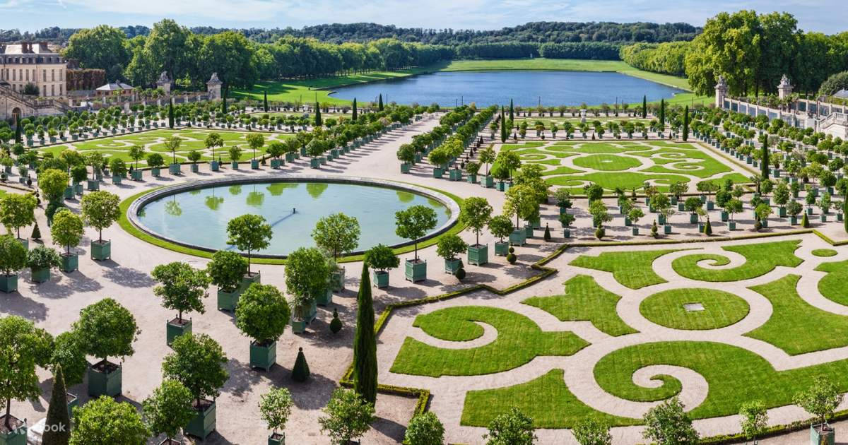 凡爾賽宮花園「Parterres」的壯麗全景 壁紙 — 從高空俯瞰一座宏偉的正式花園，擁有對稱的綠色草坪、幾何圖案、圓形池塘和遠處的湖泊，周圍環繞著茂密的樹木，陽光明媚。
