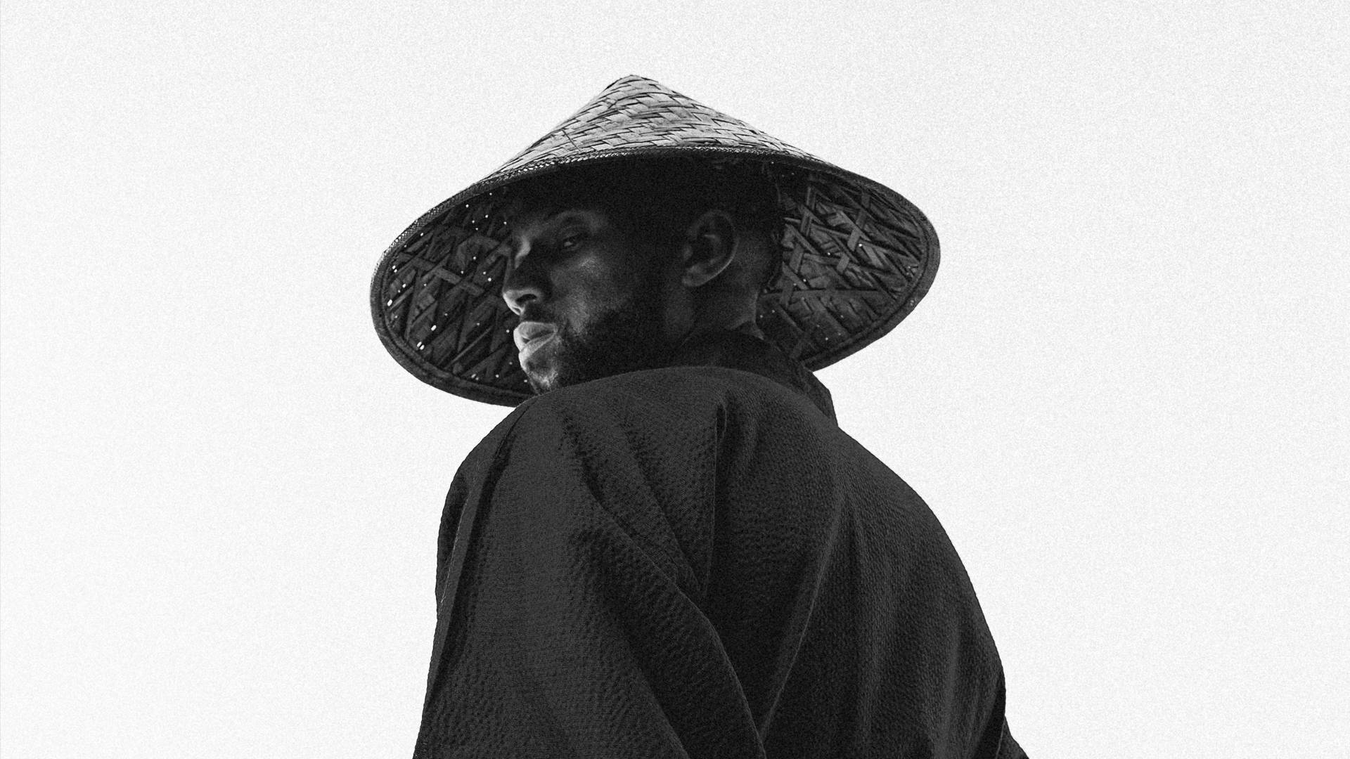 凱利恩·阿科斯塔黑白藝術照 壁紙 — 一個戴著編織錐形帽和深色長袍的男人，在純白背景上轉頭越過肩膀。