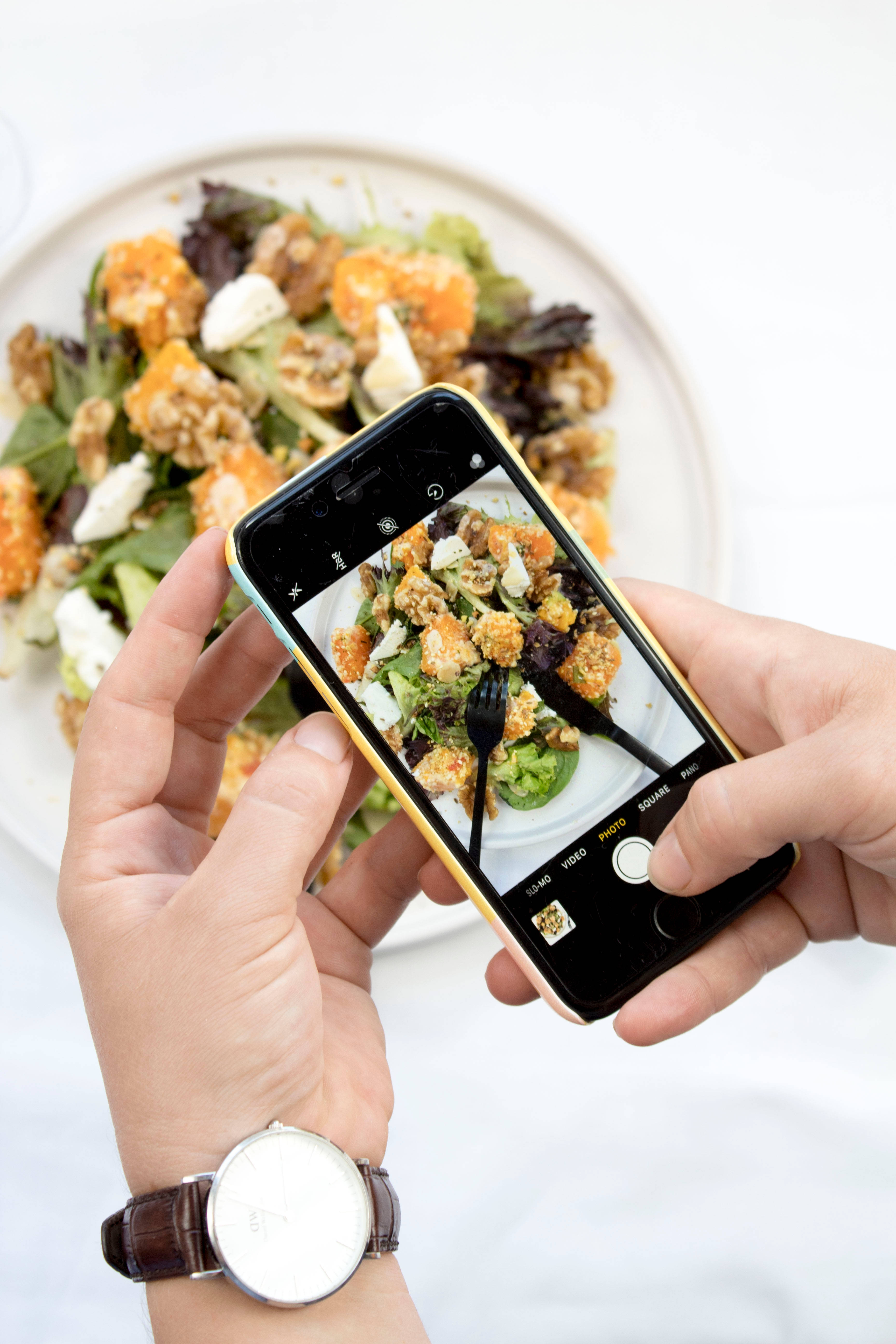 凱撒沙拉美食 iPhone 壁紙 — 雙手握住智能手機，拍攝白色碟上色彩繽紛的沙律，配有麵包丁、芝士和蔬菜。
