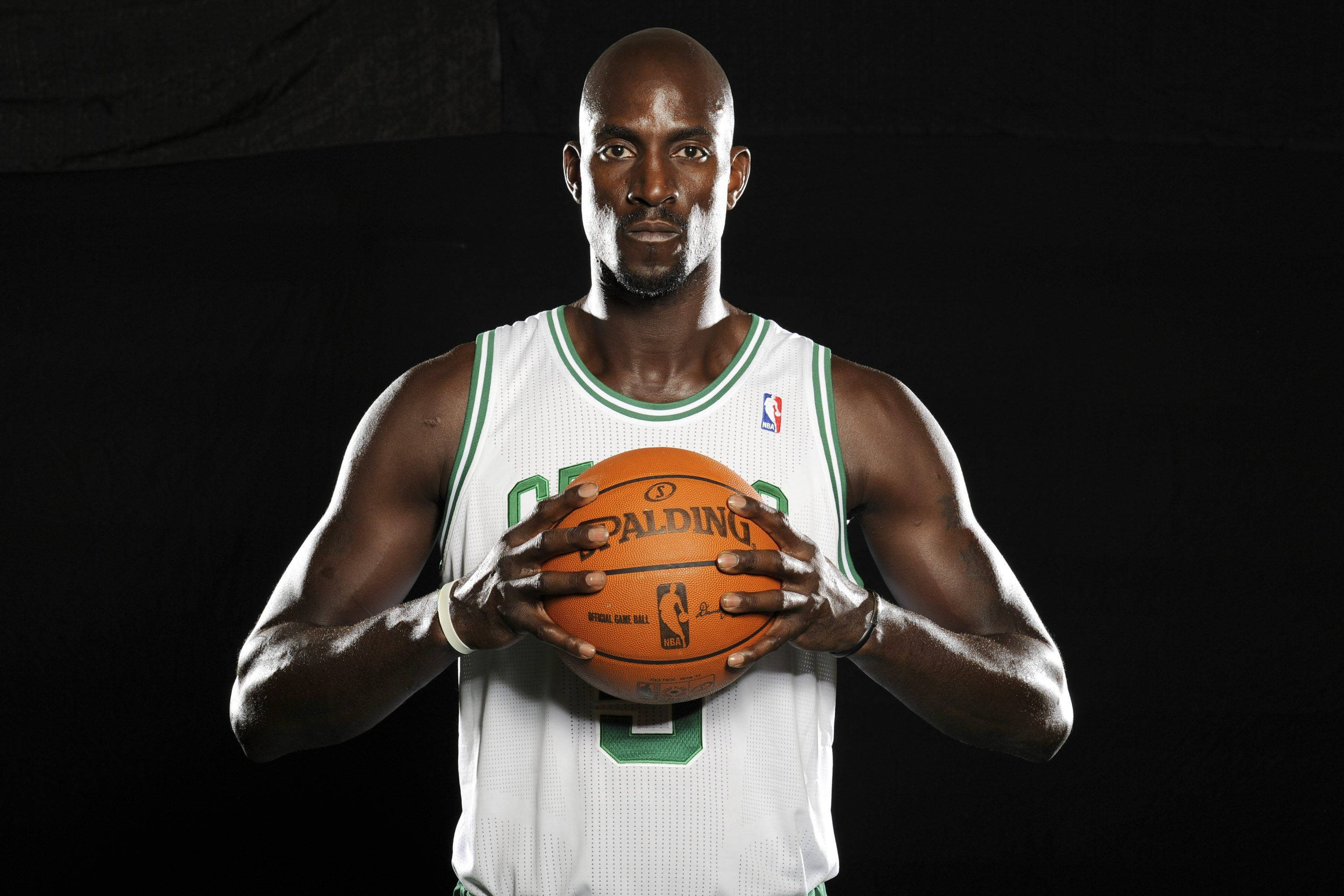 凱文·加內特 NBA 勞倫斯球員 壁紙 — 一位光頭黑人籃球運動員身穿白色和綠色球衣，在黑色背景前拿著一個橙色 Spalding 籃球。