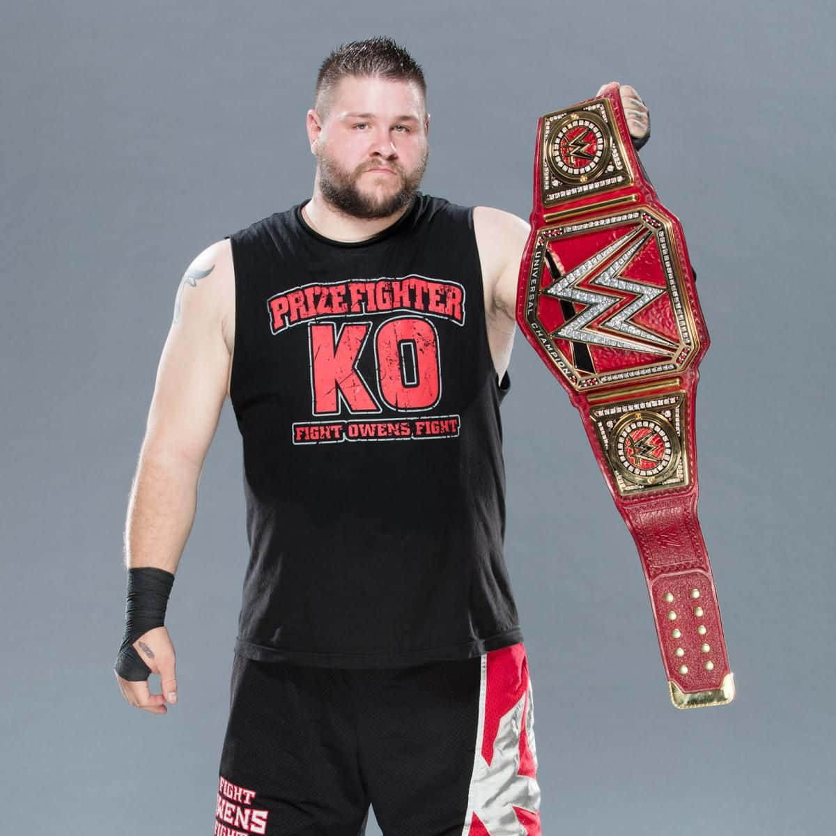 凱文·歐文斯 WWE 全球冠軍腰帶壁紙 壁紙 — 一位留鬍鬚的肌肉發達摔角手，身穿印有紅色文字的黑色背心，手持紅金冠軍腰帶。