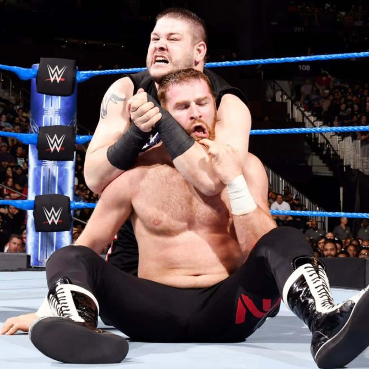 凱文·歐文斯 與薩米·扎恩 WWE 比賽 壁紙 — 兩名肌肉發達的摔角手在燈光明亮的擂台上激烈纏鬥，擂台有藍色繩索和WWE標誌，觀眾在觀看。