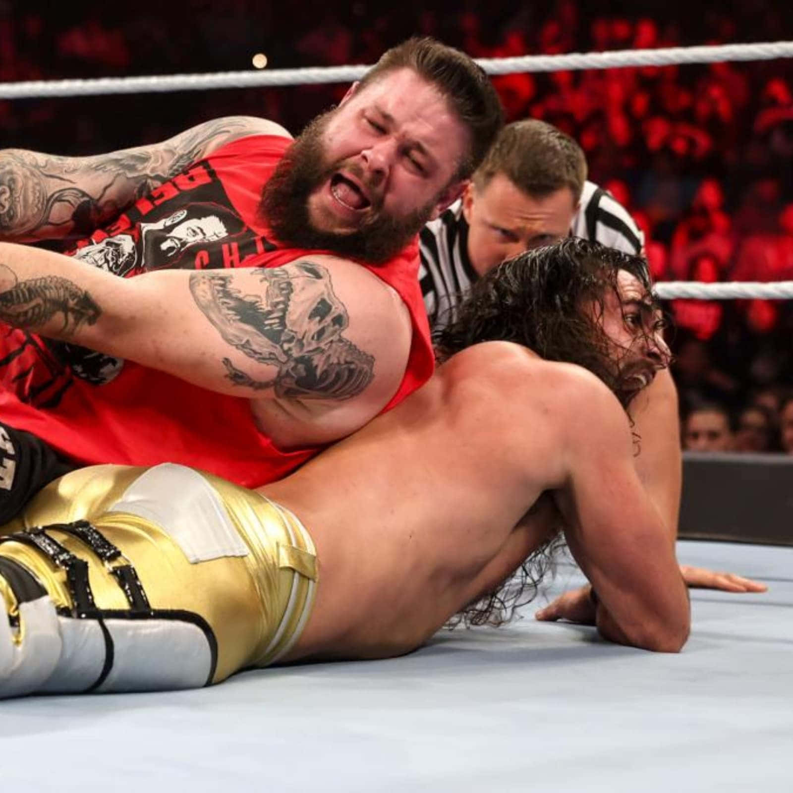 凱文·歐文斯 與賽斯·羅林斯 WWE 壁紙 — 一位留鬍鬚的摔角手身穿紅色襯衫，在明亮的場館燈光下，於一場激烈的職業摔角比賽中與身穿金色短褲的對手扭打。