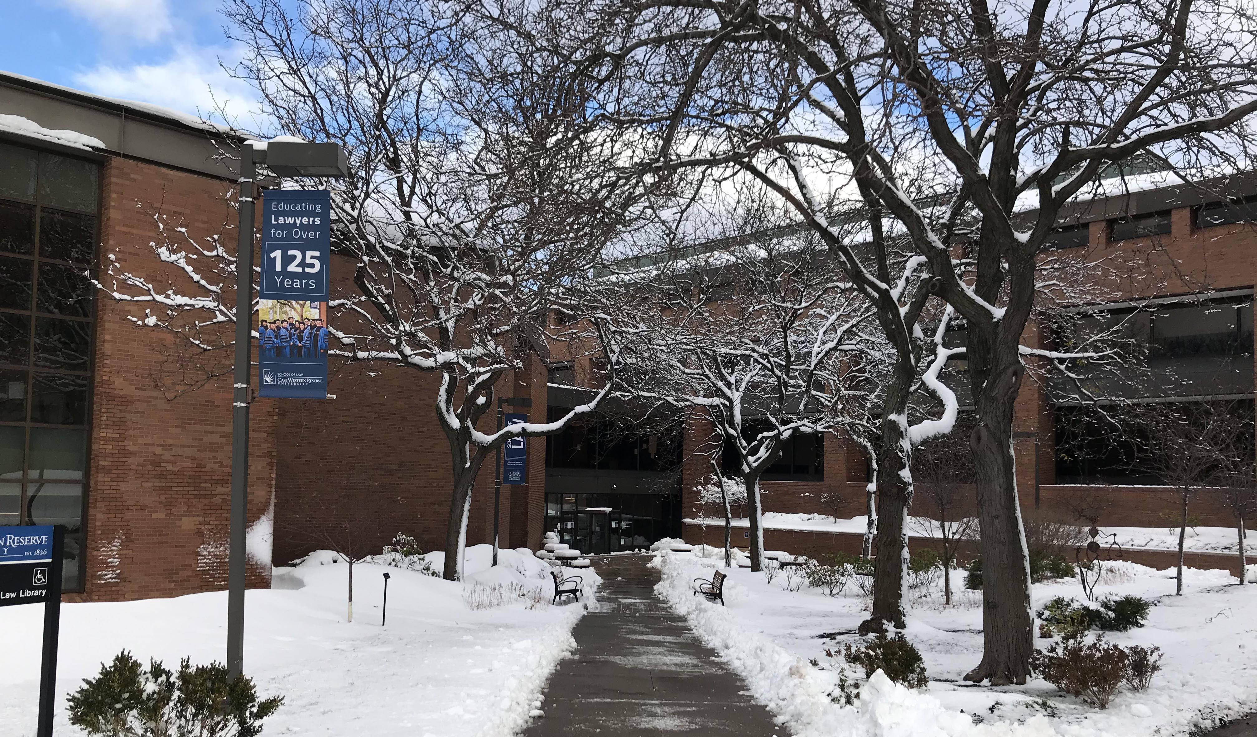 凱斯西儲大學冬季風景 壁紙 — 白雪覆蓋著一條寧靜的校園小徑，兩旁是光禿禿的樹木和磚砌建築，天空部分多雲的冬日天空。