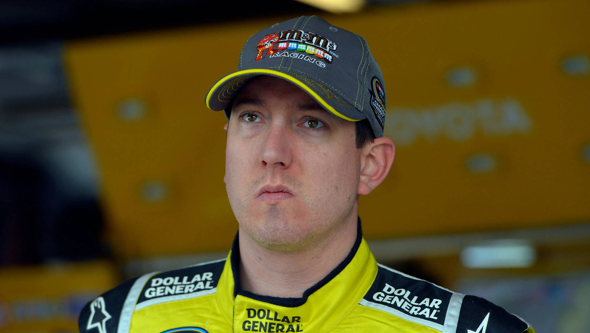 凱爾·布希無表情凝視 壁紙 — 一位NASCAR車手身穿黃黑賽車服的近距離特寫，神情專注。